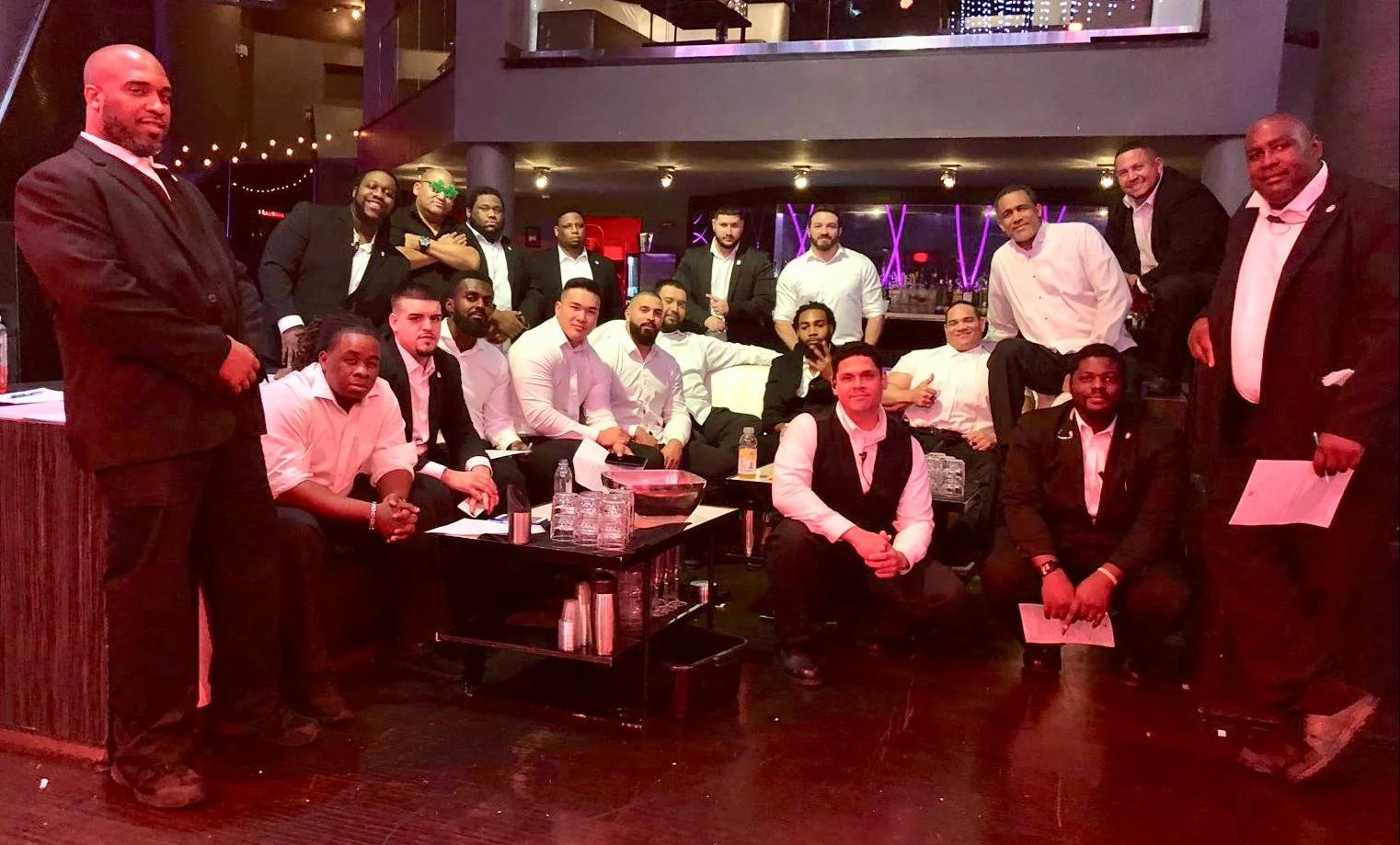 Group of men dressed in formal attire gathered in a modern indoor setting with dim lighting and purple neon lights in the background.