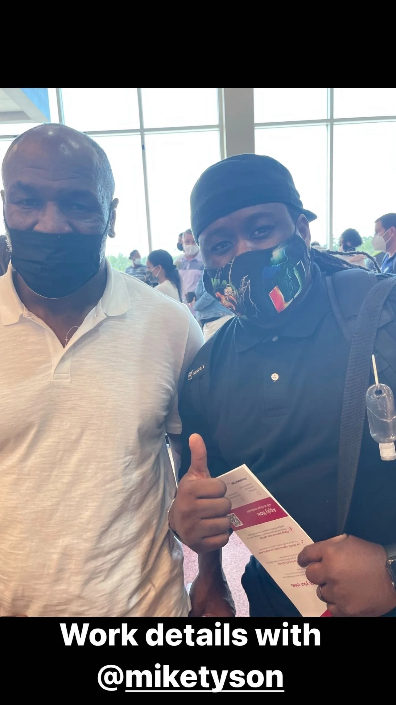 Two men wearing masks at an indoor event, one holding a ticket or document, with large windows in the background and other people in the background.