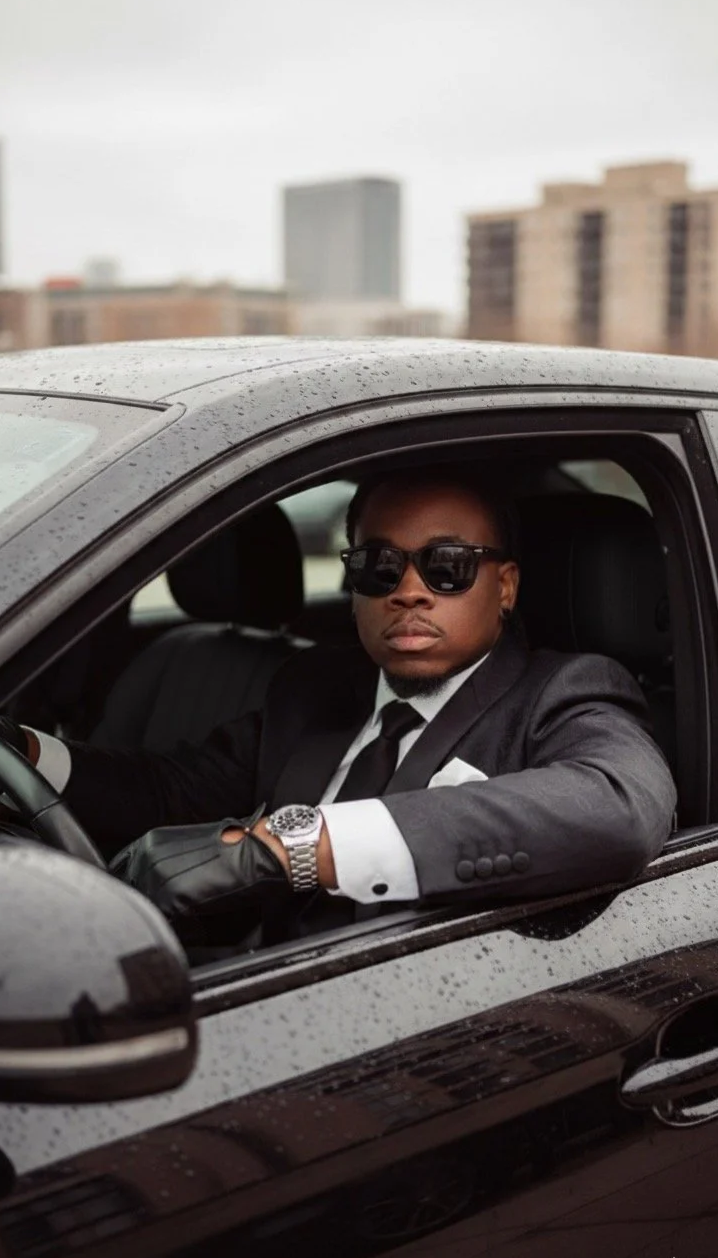 Man in a suit and sunglasses sitting in a black car with raindrops on the window, city buildings in the background.