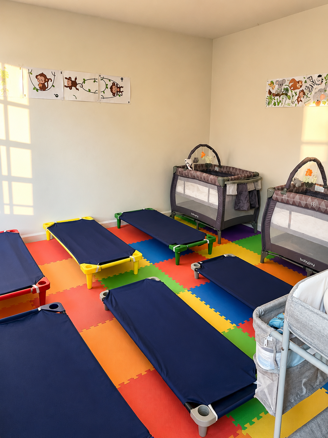 A room with colorful foam floor tiles, several small blue beds, two portable cribs, and a changing table.