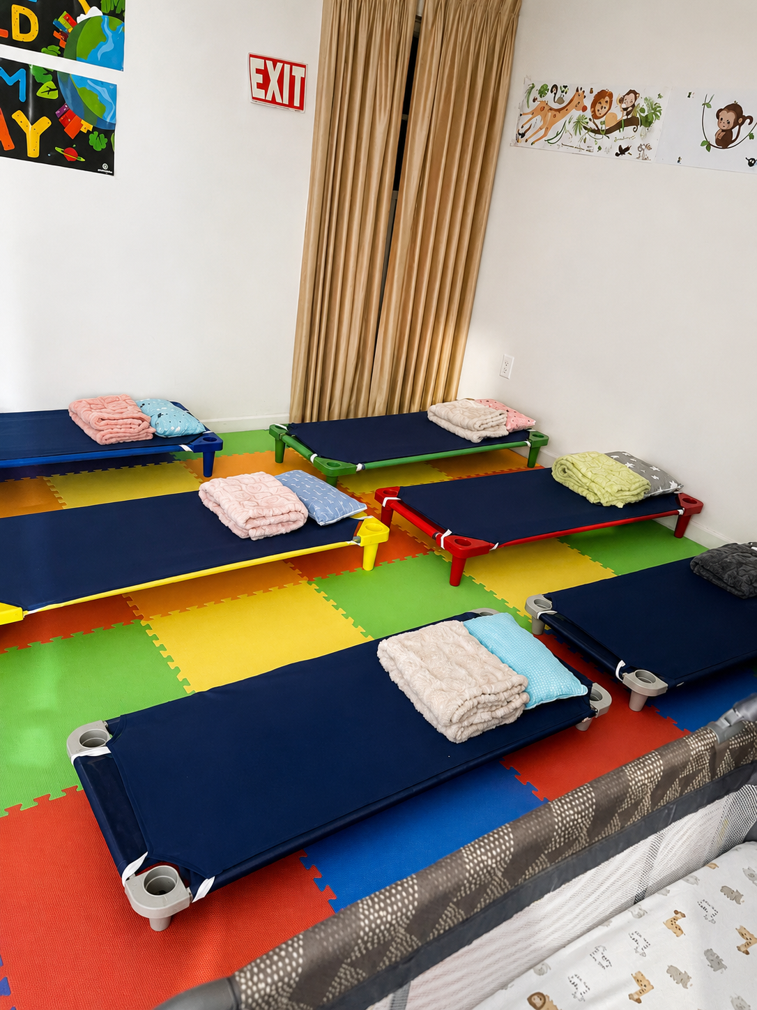 A room with colorful foam mats on the floor, containing small nurse-like beds with folded towels and pillows, set up for children to rest or nap.