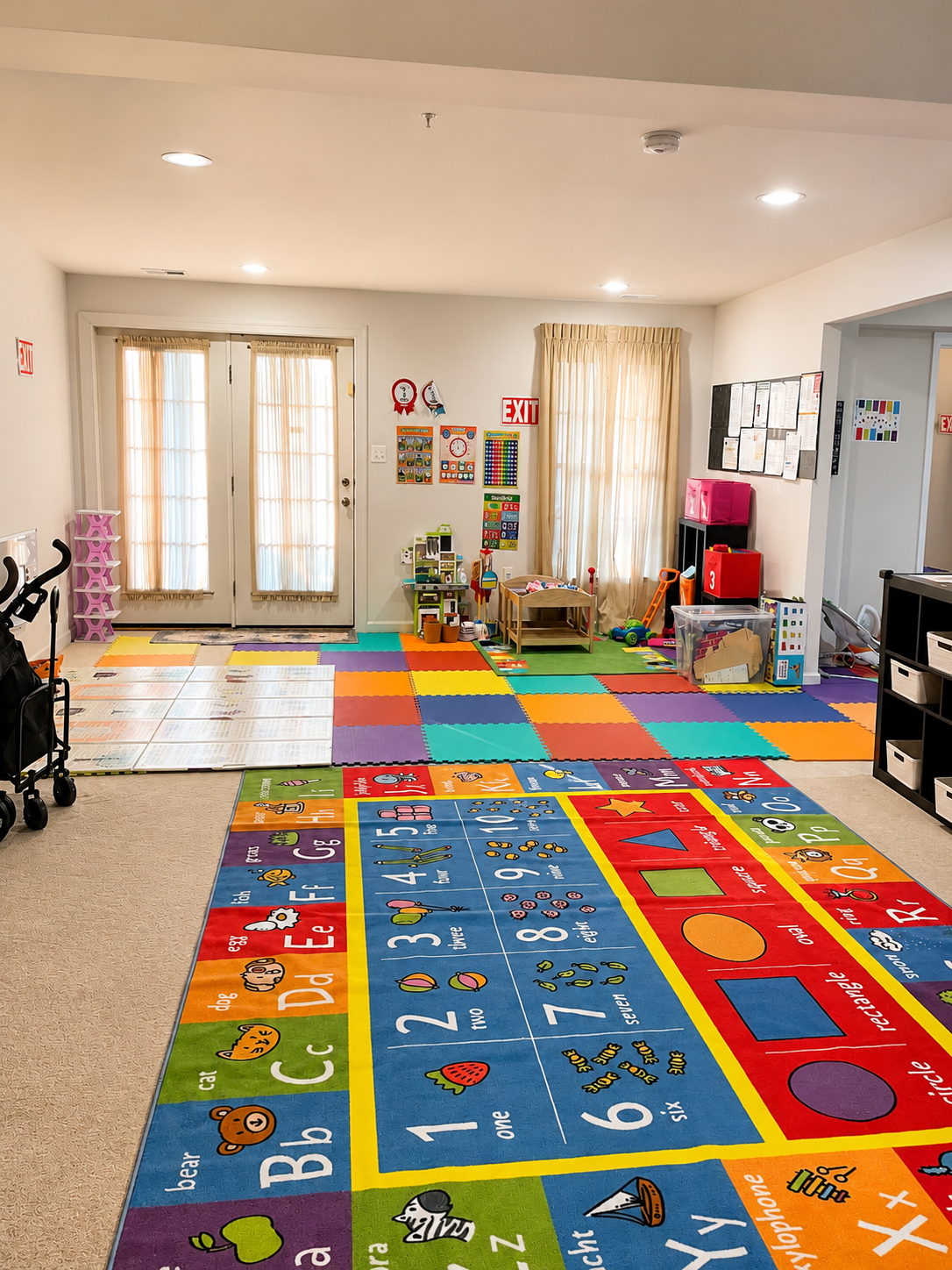 Colorful children's playroom with a bright alphabet and number rug, foam puzzle floor tiles, toys, and storage bins. Double glass doors allow natural light, with beige curtains, ceiling lights, and children's educational decorations on the walls.