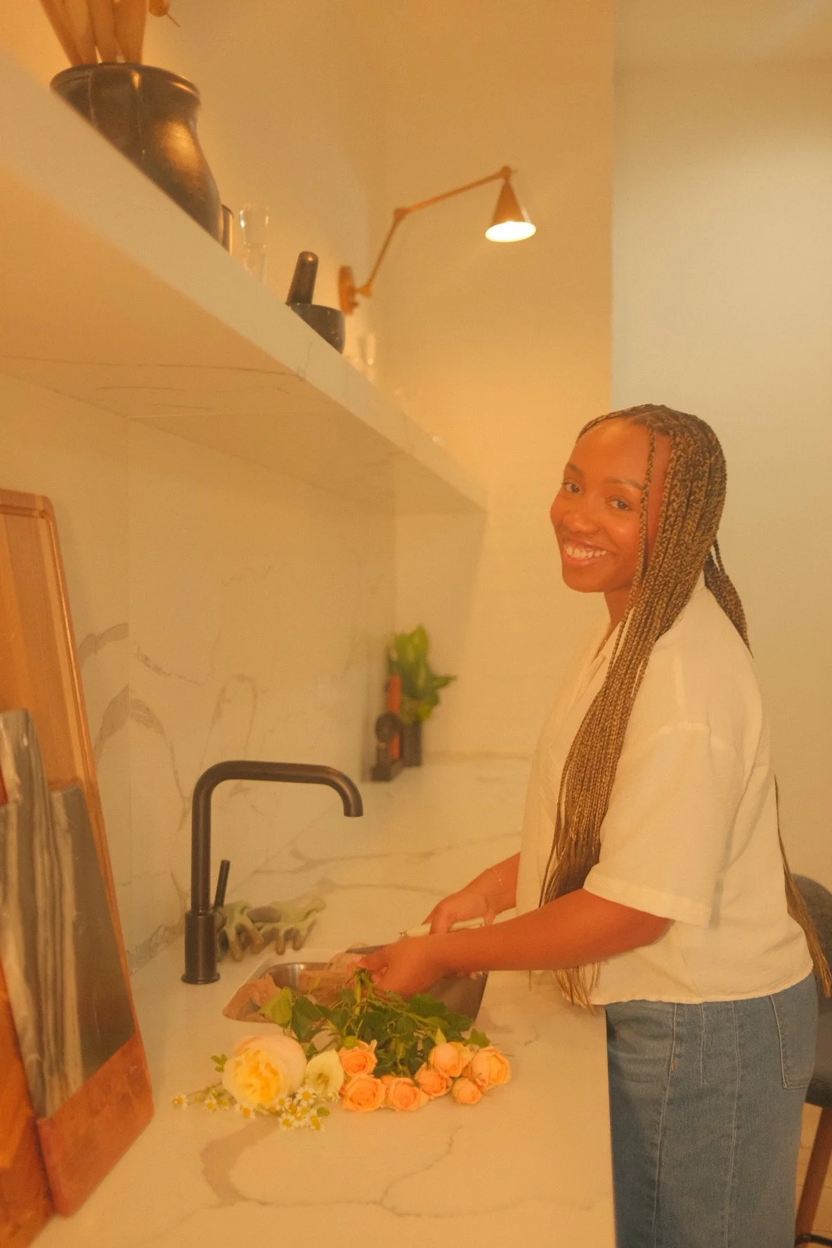 A woman with braided hair smiling as she cuts stems at a kitchen sink. There are peach roses on the counter nearby and a plant in the background.