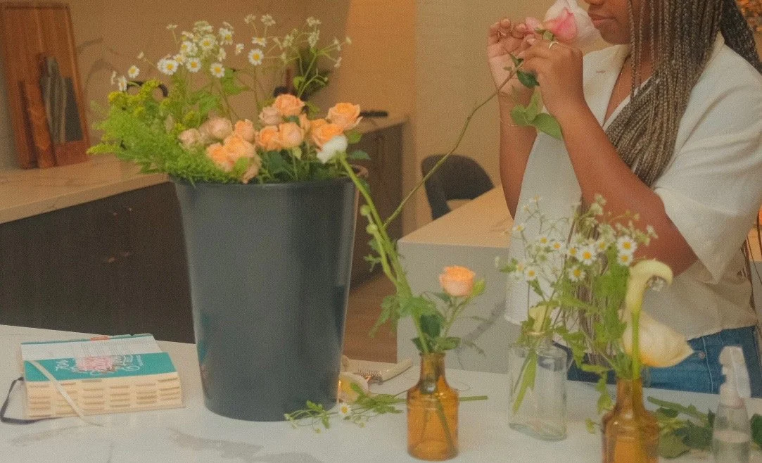 A woman arranging flowers on a table with vases and flowers, including caila lilies and roses, in a room with a counter and a Bible in the background.