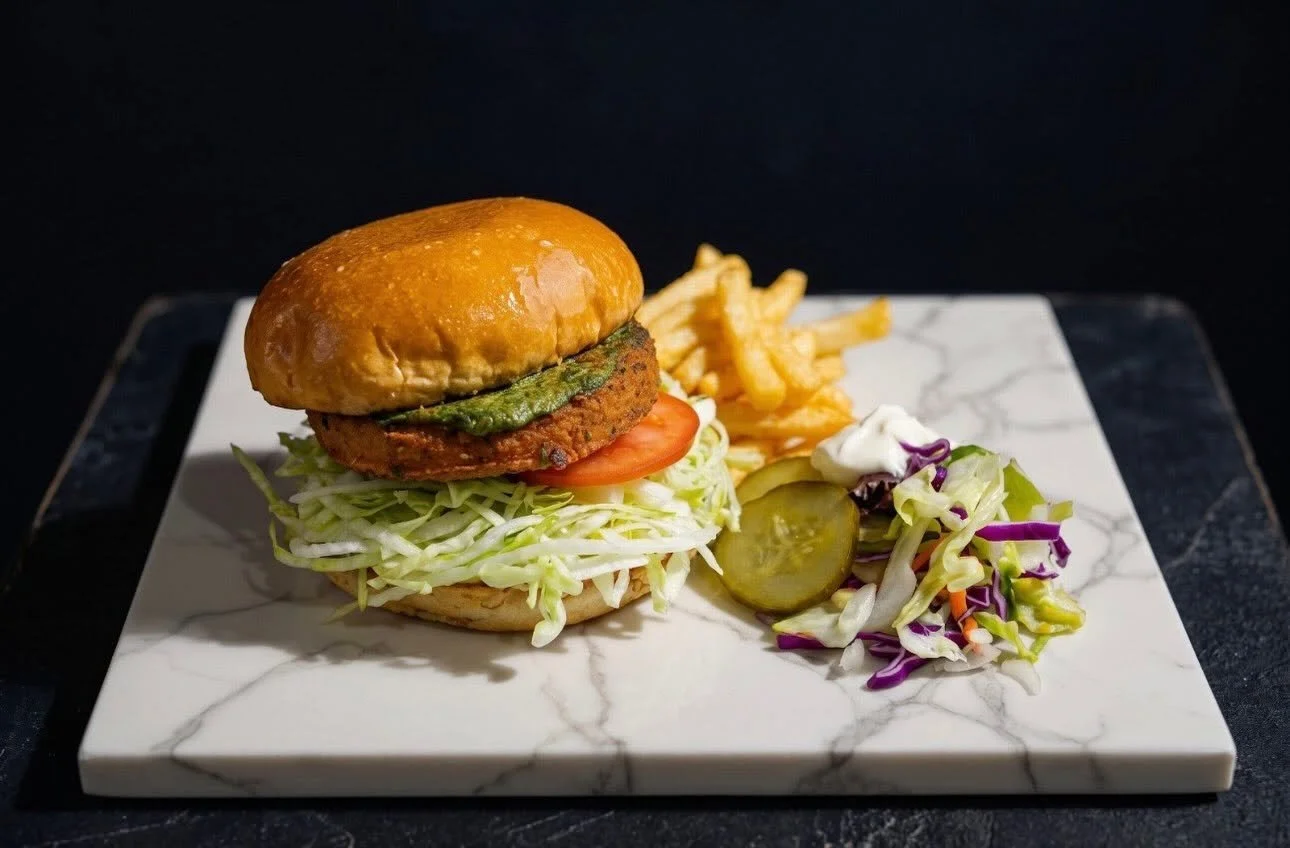 A burger with lettuce, tomato, and fried patty topped with pickles, served with fries and a side salad on a white marble plate.