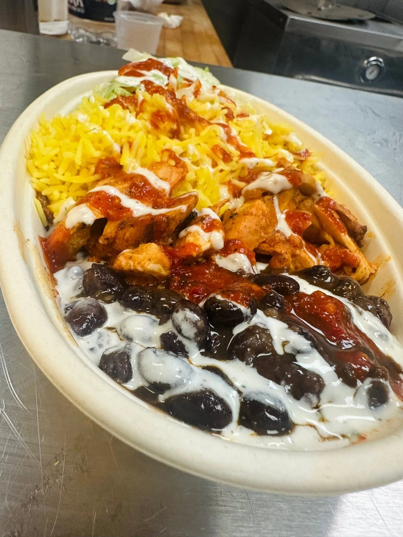 A white oval plate containing cooked rice, fried chicken with sauce, black beans, and shredded lettuce with drizzled sauces, on a stainless steel surface.
