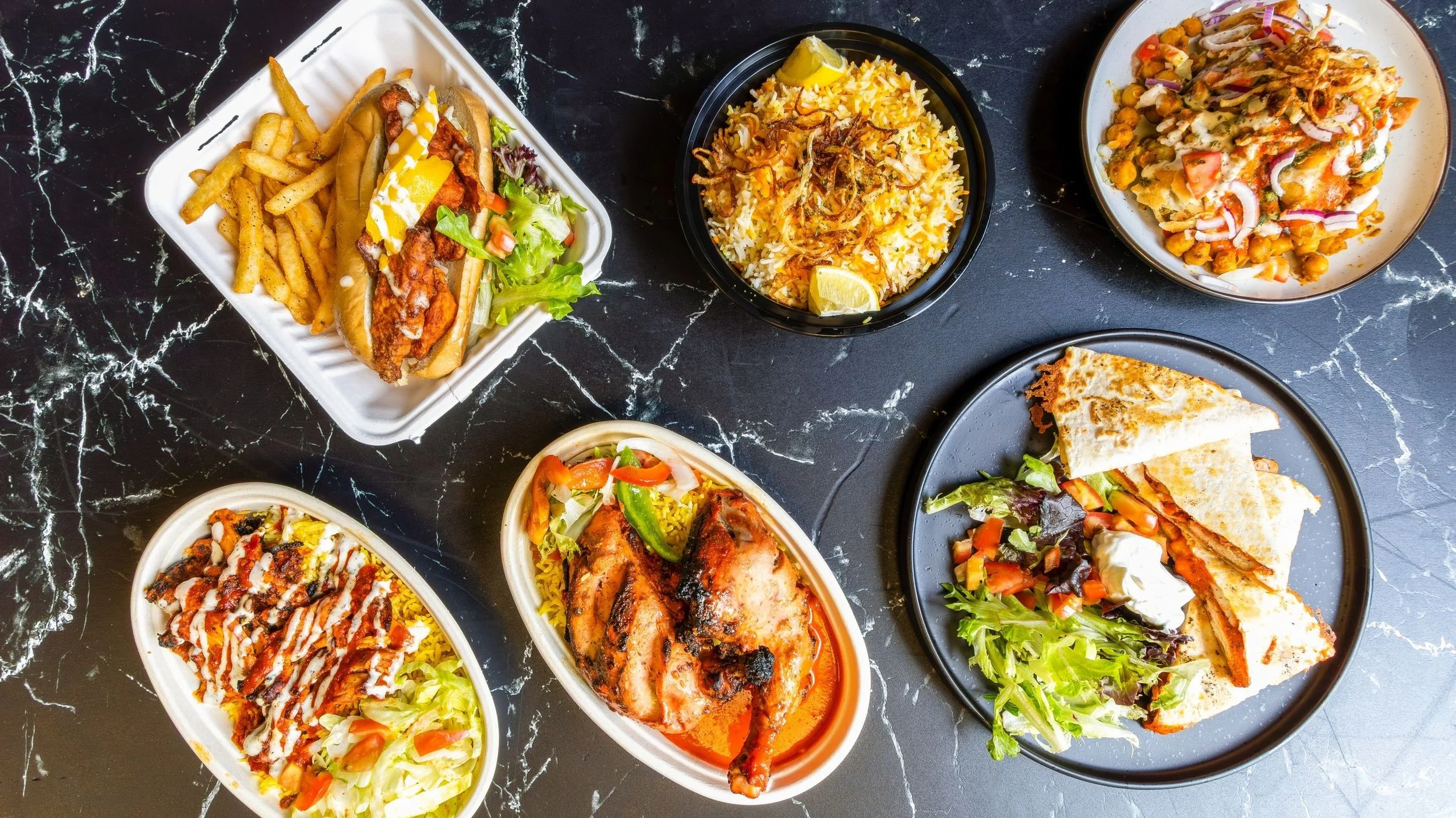 Assorted Indian dishes and fast food on black marble surface, includes biryani, grilled chicken, naan, salad, samosas, and a hot dog with fries, all in bowls and plates.