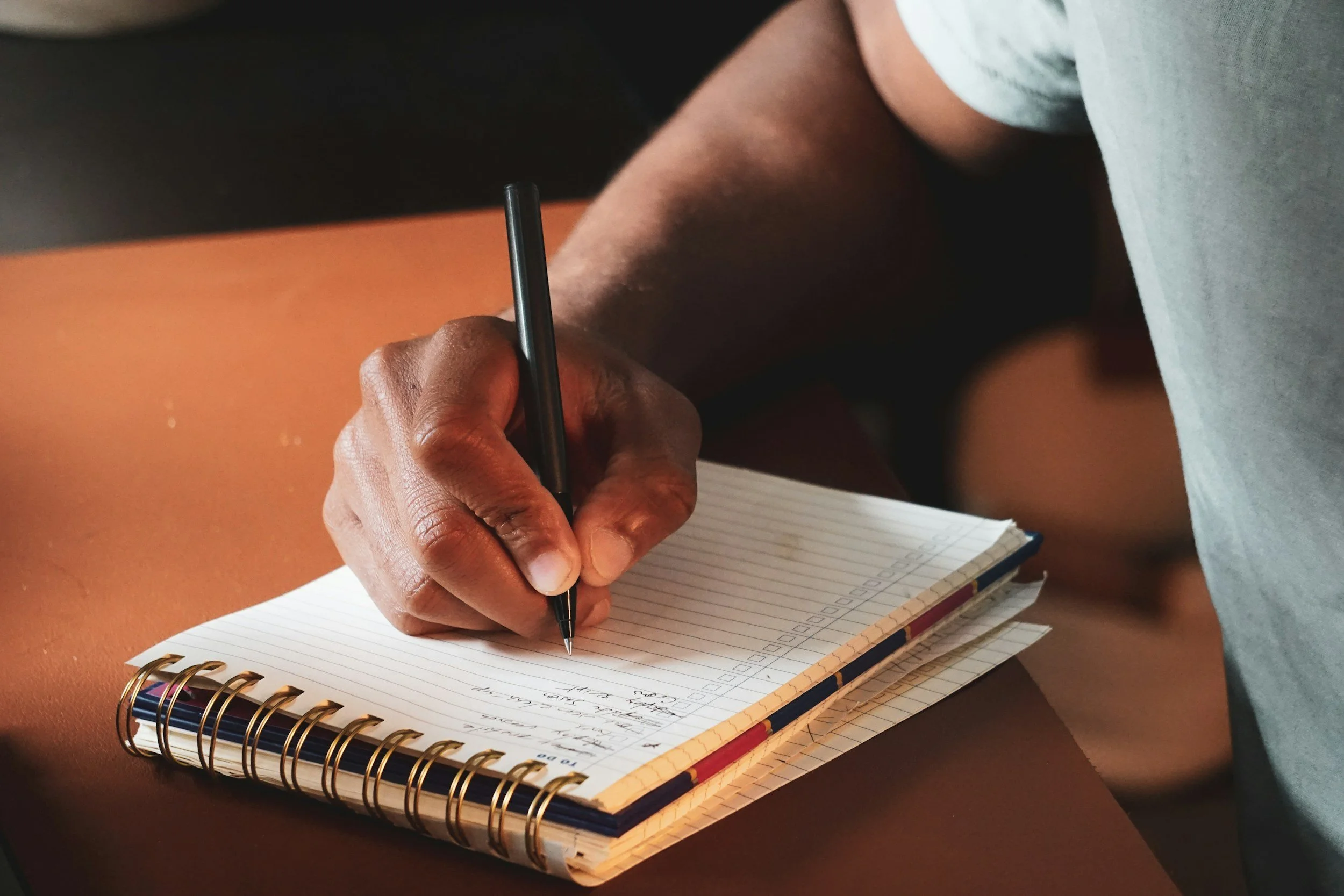 A person with dark skin is writing in a spiral notebook with ruled pages using a black pen, resting on a brown surface.