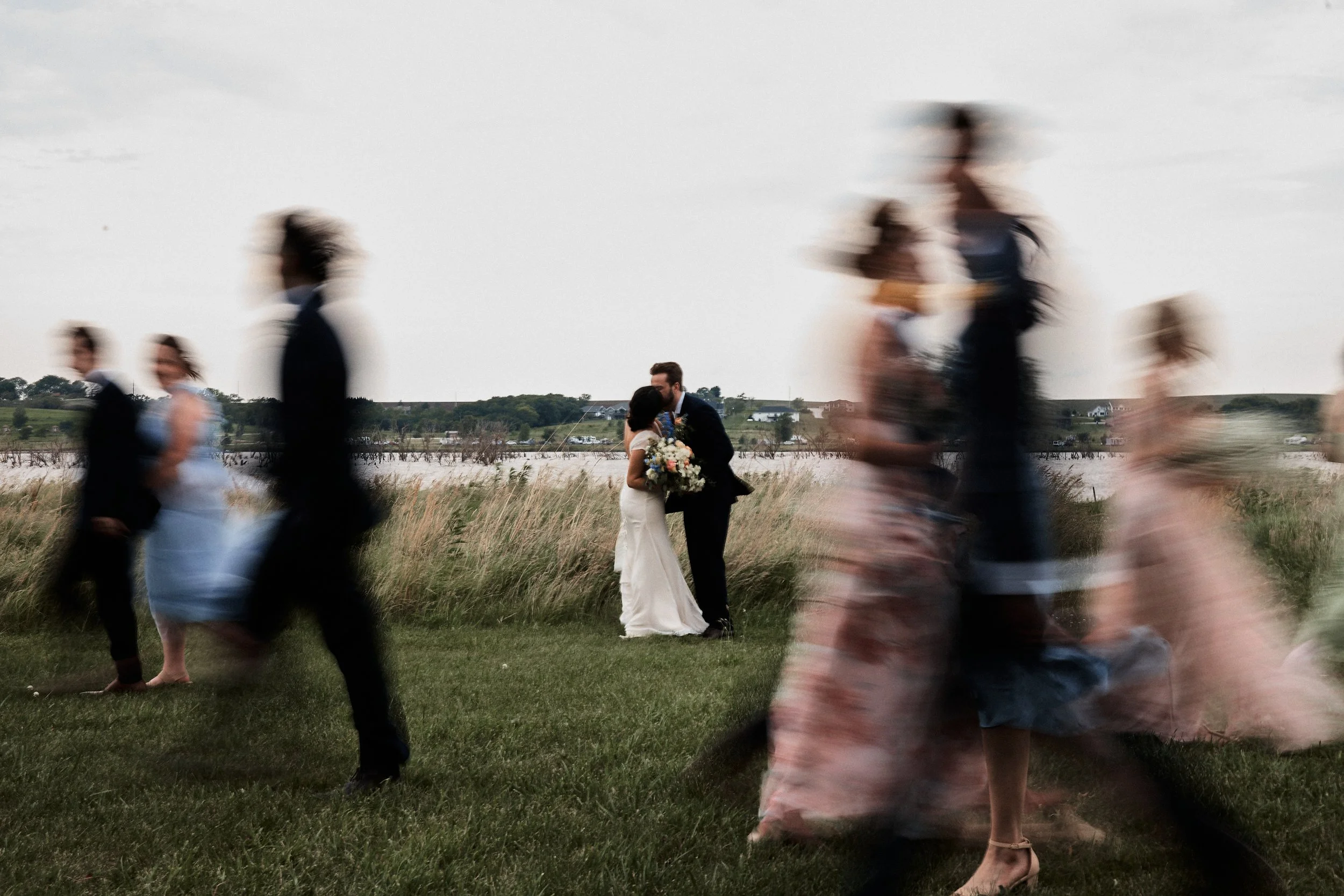 A couple, bride and groom, kissing in a field during sunset while multiple guests walk by in motion blur.