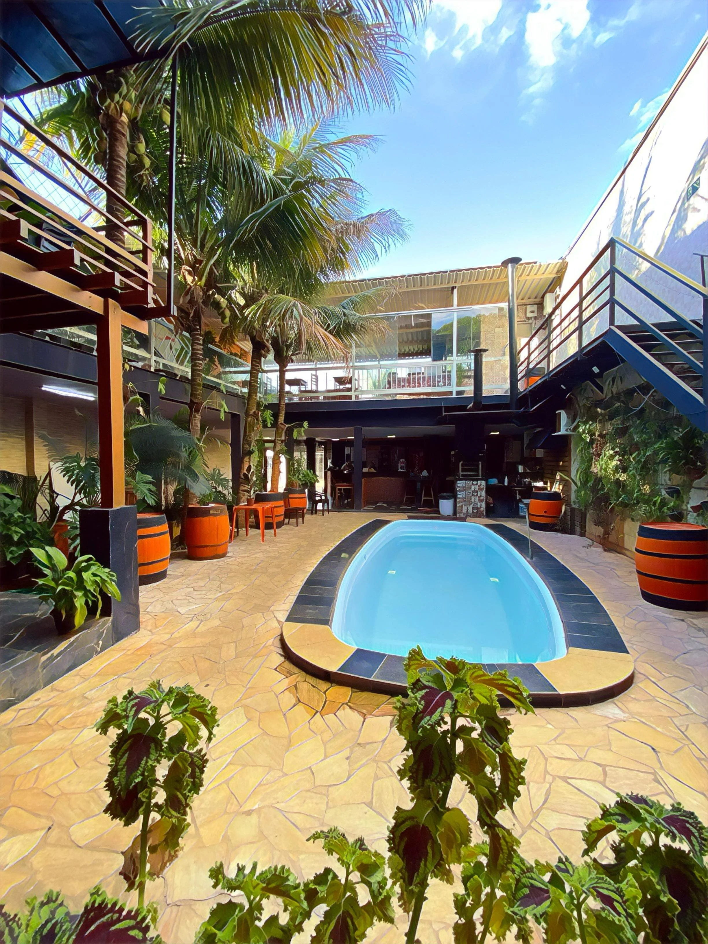 Piscina em um espaço fechado com muitas plantas verdes, incluindo palmeiras, e móveis ao redor, sob um teto de vidro com céu azul visível.