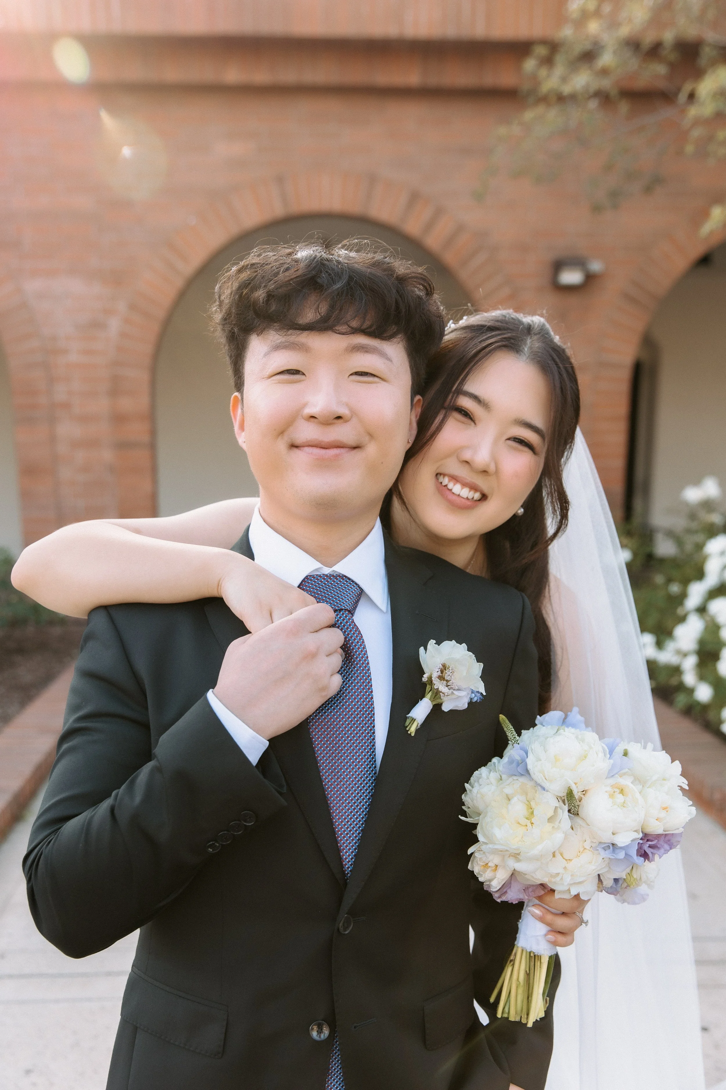 A newlywed couple, a man in a black suit and a woman in a wedding dress, standing outdoors in front of a brick building, smiling and posing for the photo, with the woman holding a bouquet of white and light purple flowers.