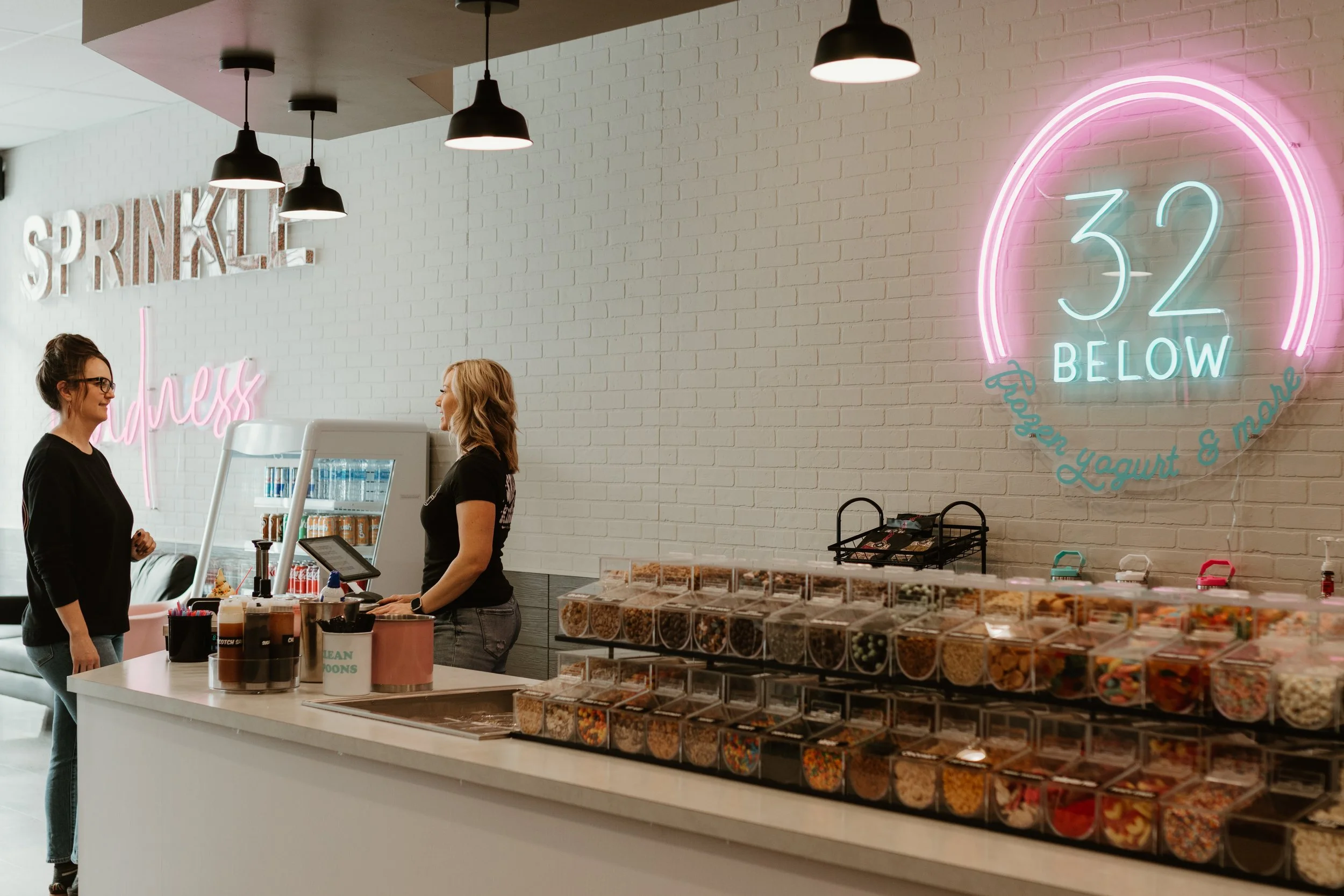 Inside a modern ice cream or frozen yogurt shop, two women are standing at the counter. One woman is a customer wearing glasses and a black top, while the other is an employee wearing a black shirt. The shop has a white brick wall with neon signs that say 'SPRINKLE' and '32 BELOW.' There are various toppings in small containers on the counter, and the background features black pendant lights hanging from the ceiling.