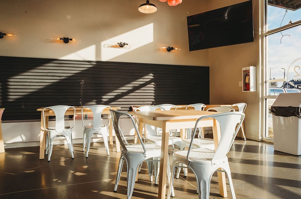 Empty restaurant with wooden tables, white chairs, a black chalkboard wall, a wall-mounted TV, and large windows letting in sunlight.