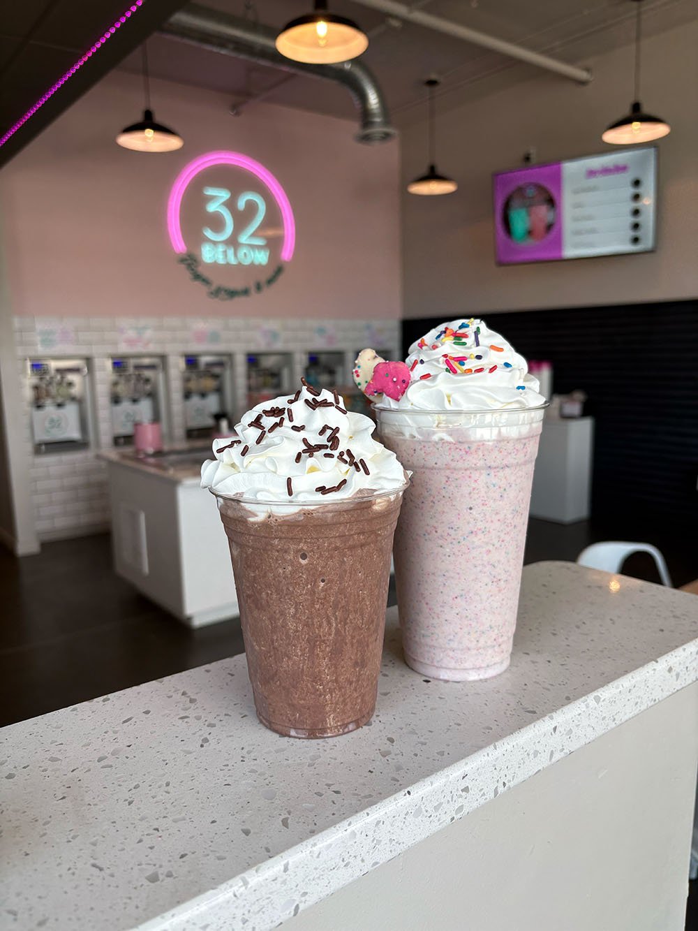 Two frosted milkshakes topped with whipped cream and sprinkles, placed on a speckled white countertop inside a shop with pink and white decor.