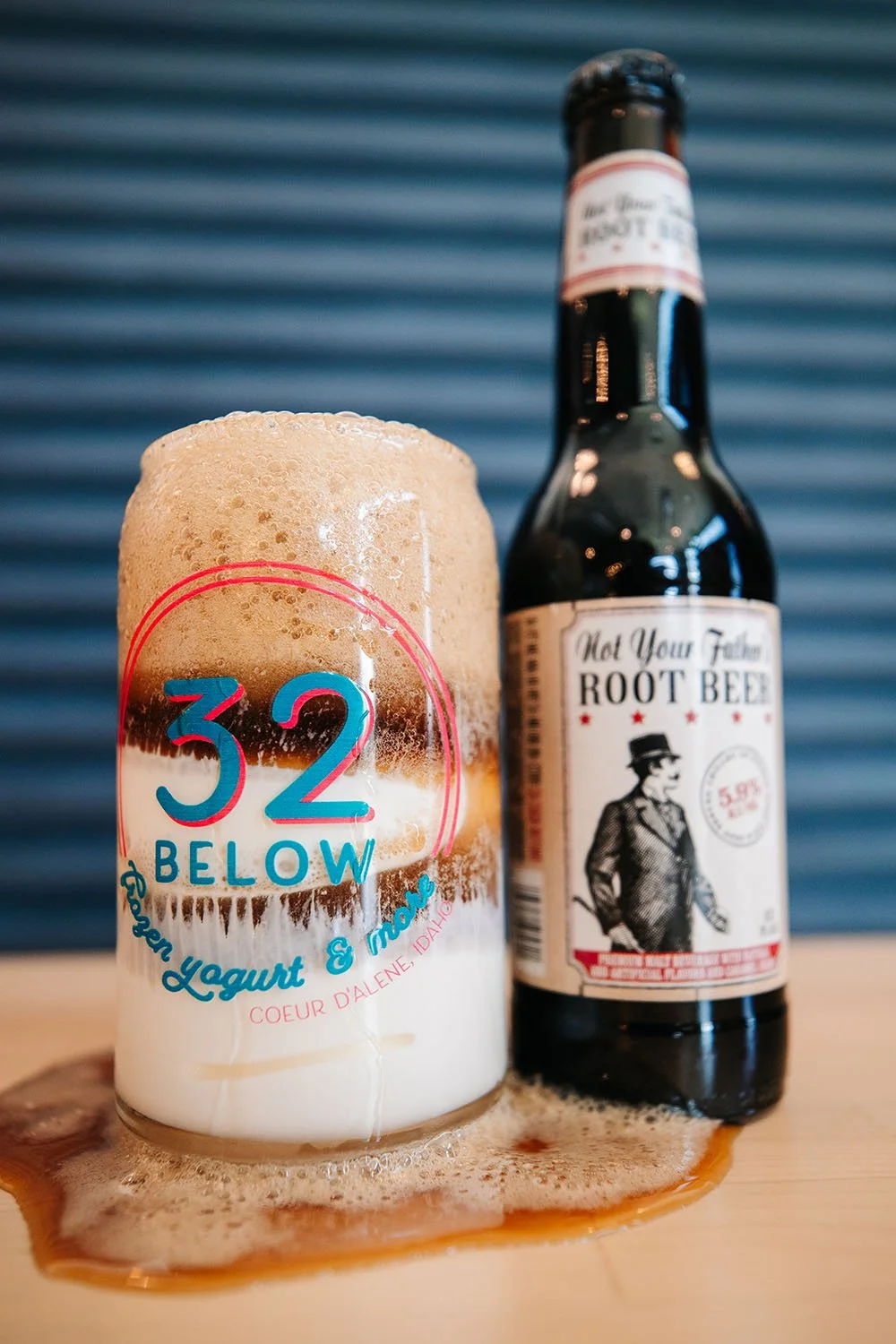 A bottle of root beer and a frothy glass of root beer with foam overflowing, sitting on a wooden surface with foam and liquid spilled around.