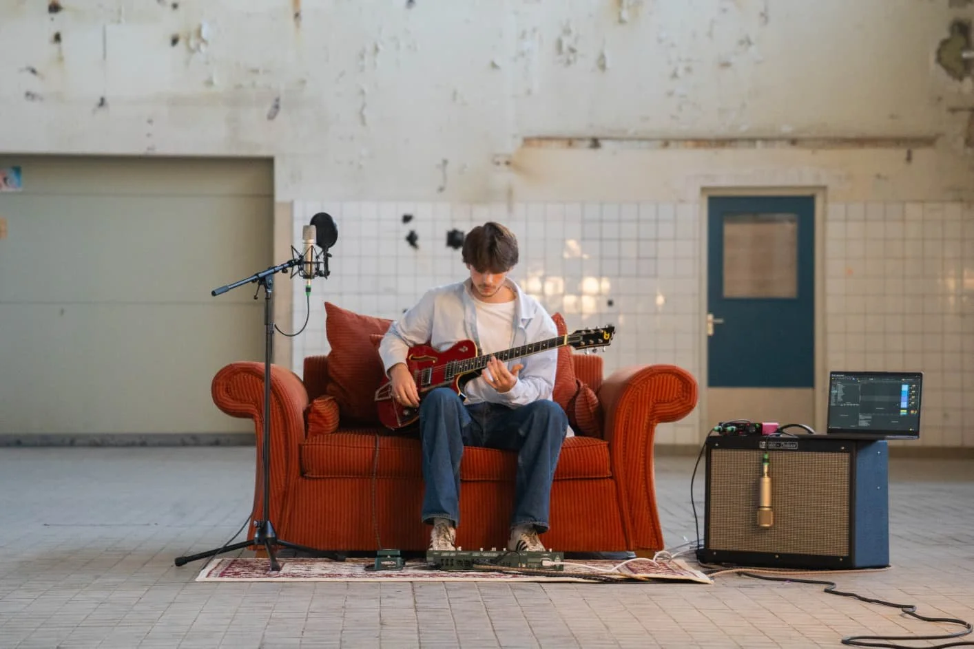A young man sitting on an orange sofa, playing an electric guitar in a room with a tiled wall, a door, music equipment, and a laptop.