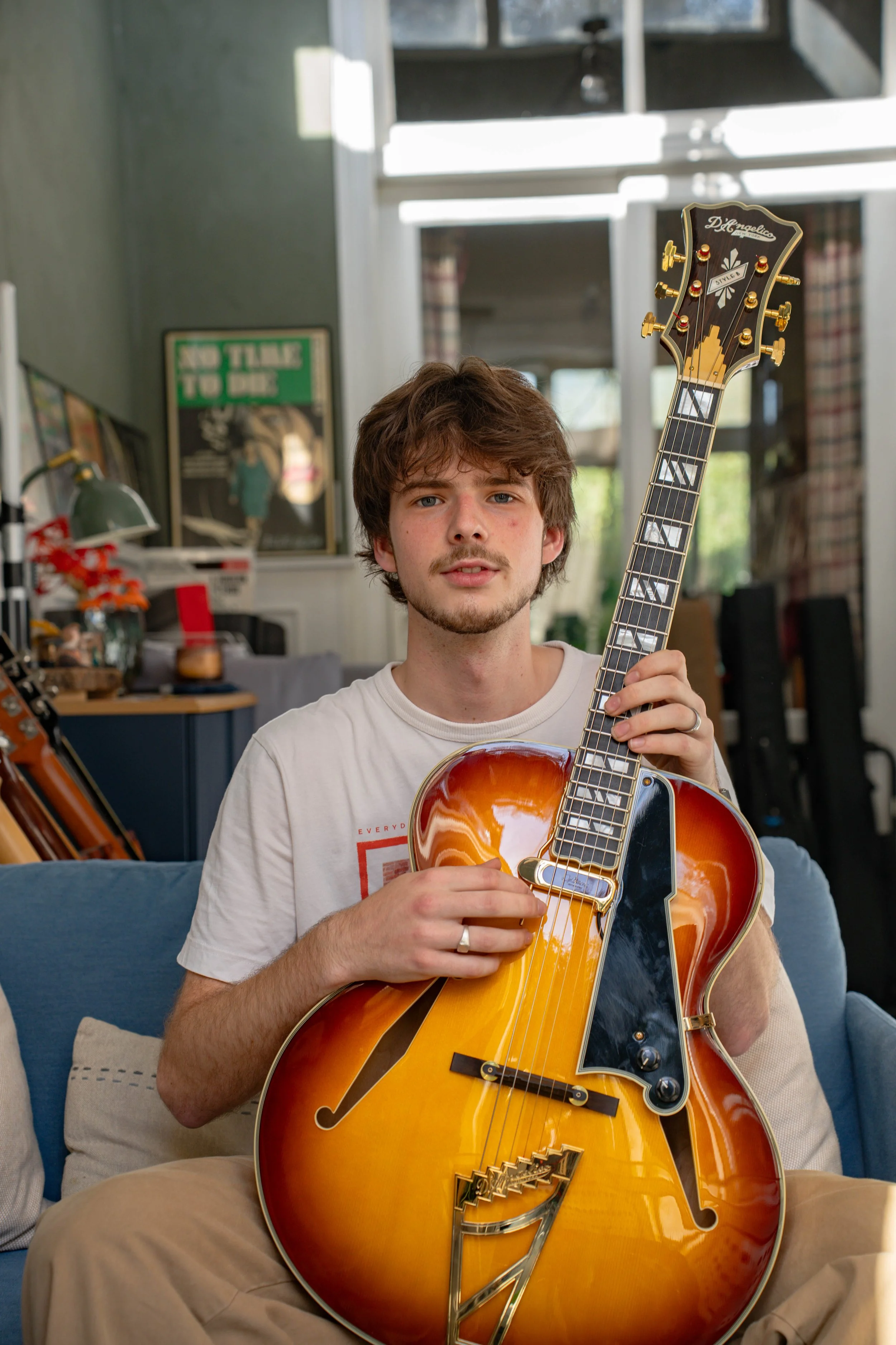 A young man with brown hair and a light beard holds an orange and black hollow body electric guitar indoors, sitting on a beige cushioned chair with a blue pillow. He wears a white t-shirt and beige pants. The background features a room with a window