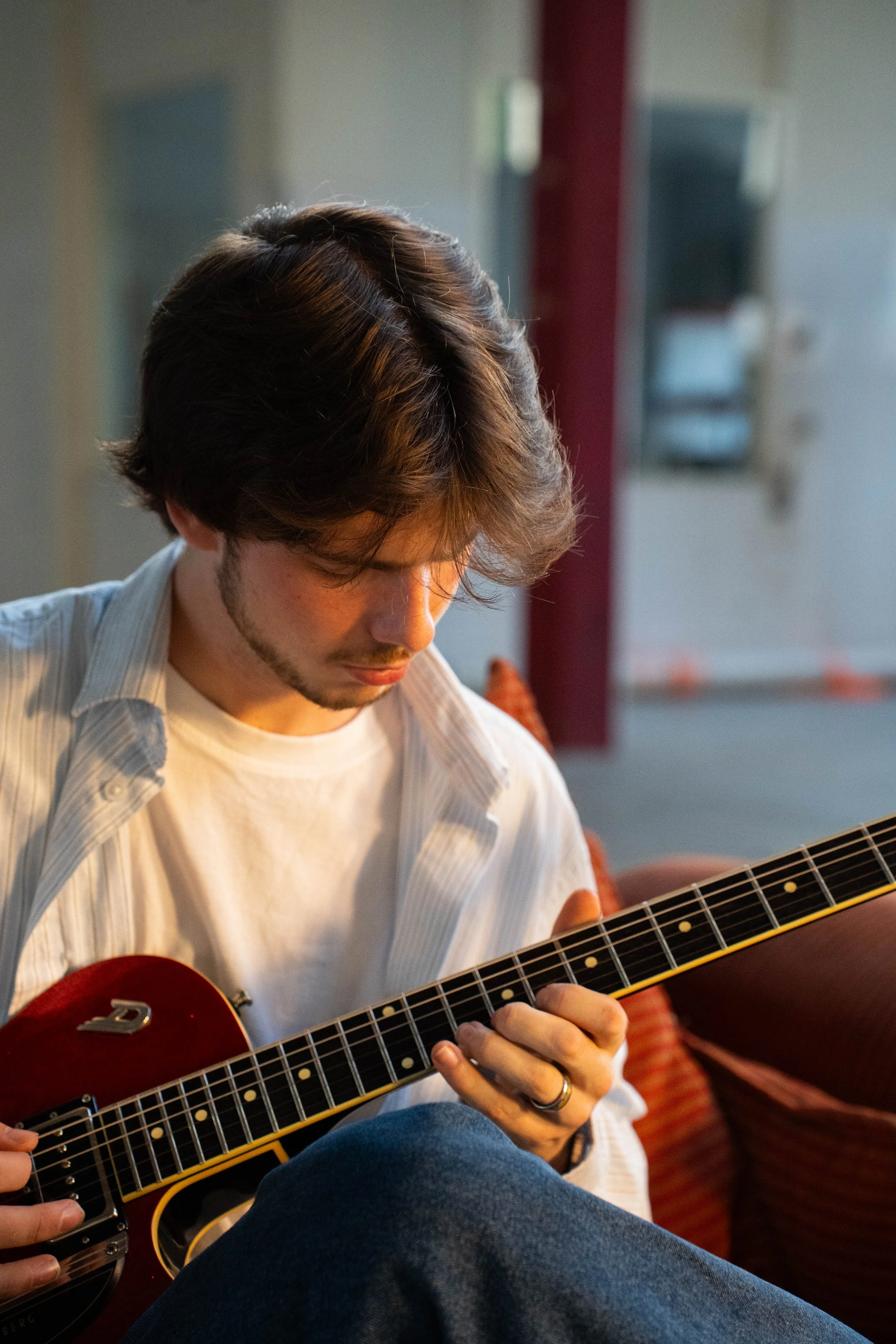 A young man with brown hair and a beard, wearing a white shirt, is sitting while playing a red acoustic guitar. The background is slightly blurred, with hints of a cozy indoor setting.
