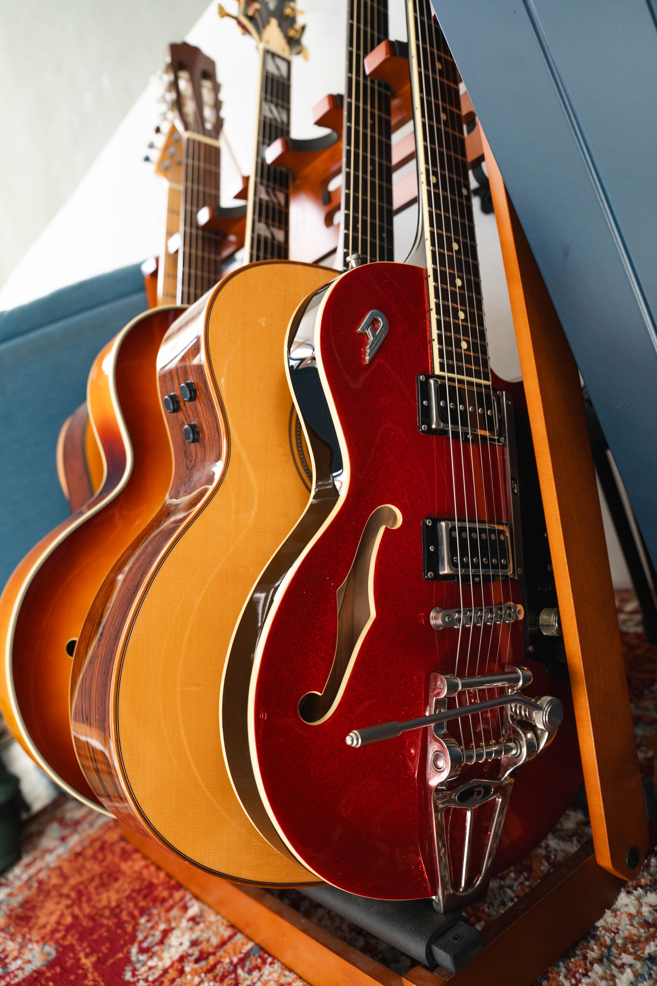 Four guitars placed on a rack with a blue piece of furniture beside them and a patterned carpet visible underneath.