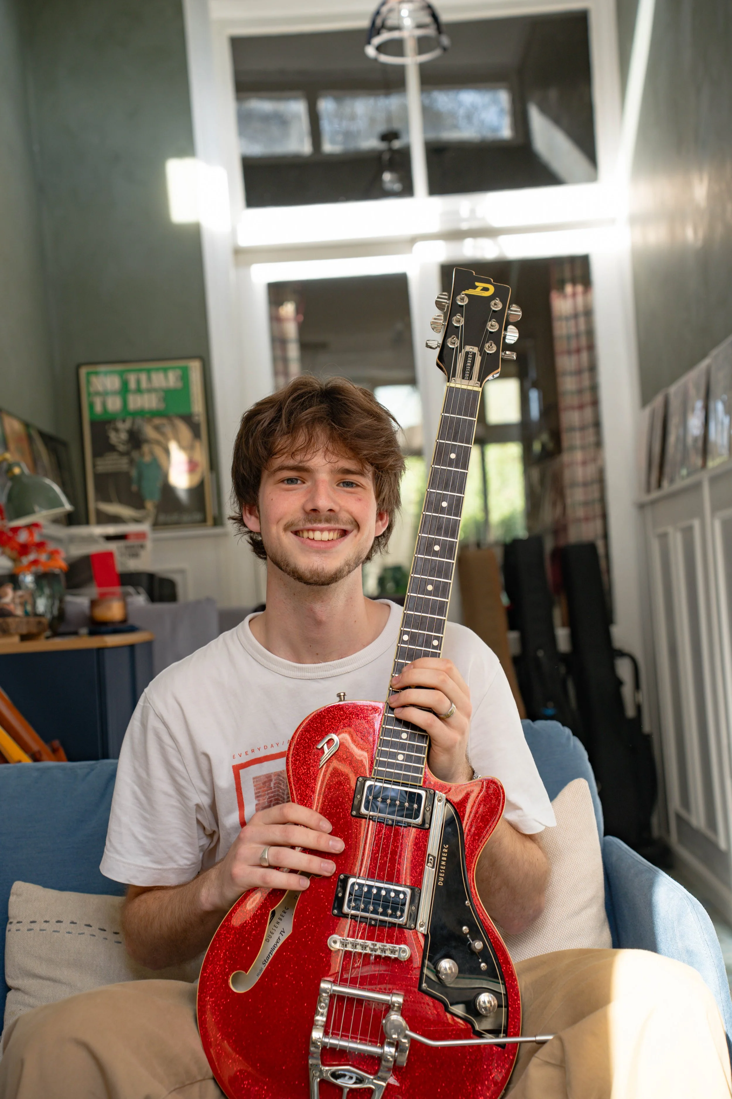 A young man with tousled brown hair and a light beard sitting on a couch, smiling, and holding a red electric guitar inside a living room with books and posters in the background.