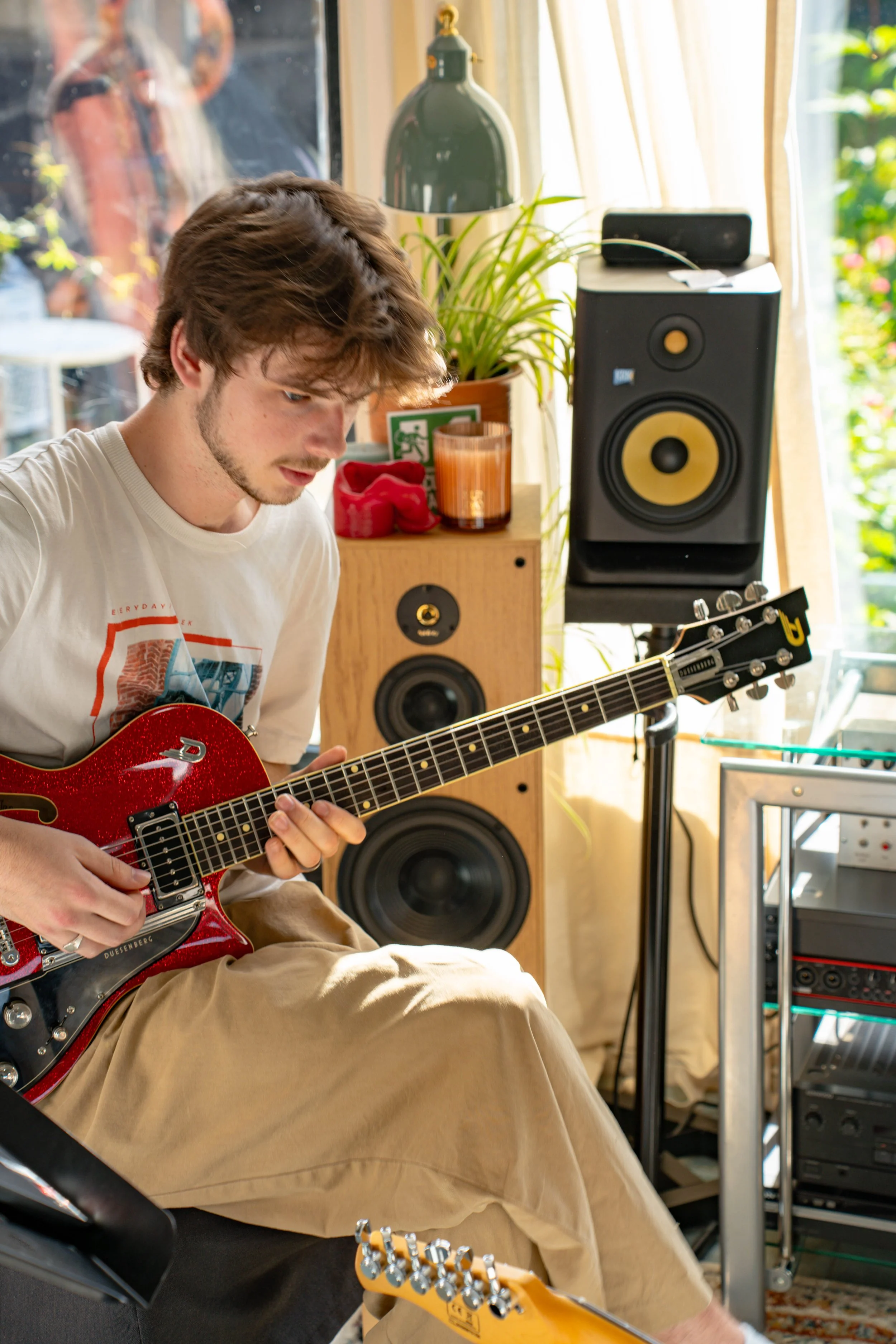 A young man with brown hair and a beard playing a red electric guitar, sitting in a bright room with large windows, green plants, and audio equipment.