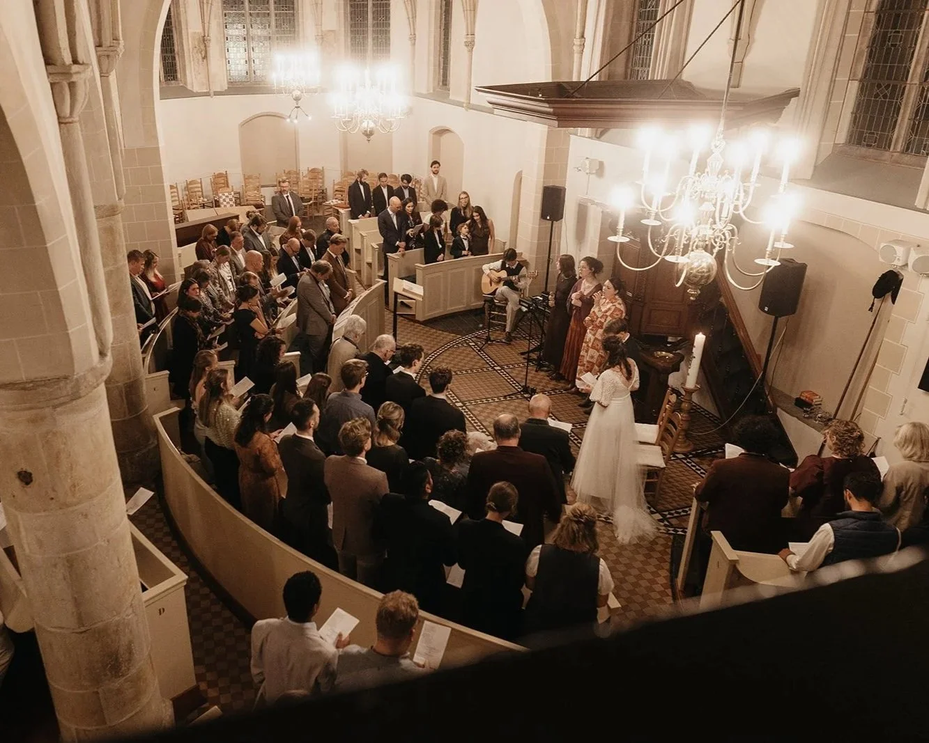A wedding ceremony taking place inside a church with a large congregation, a bride in a white dress, a guitarist, and singers performing at the front.