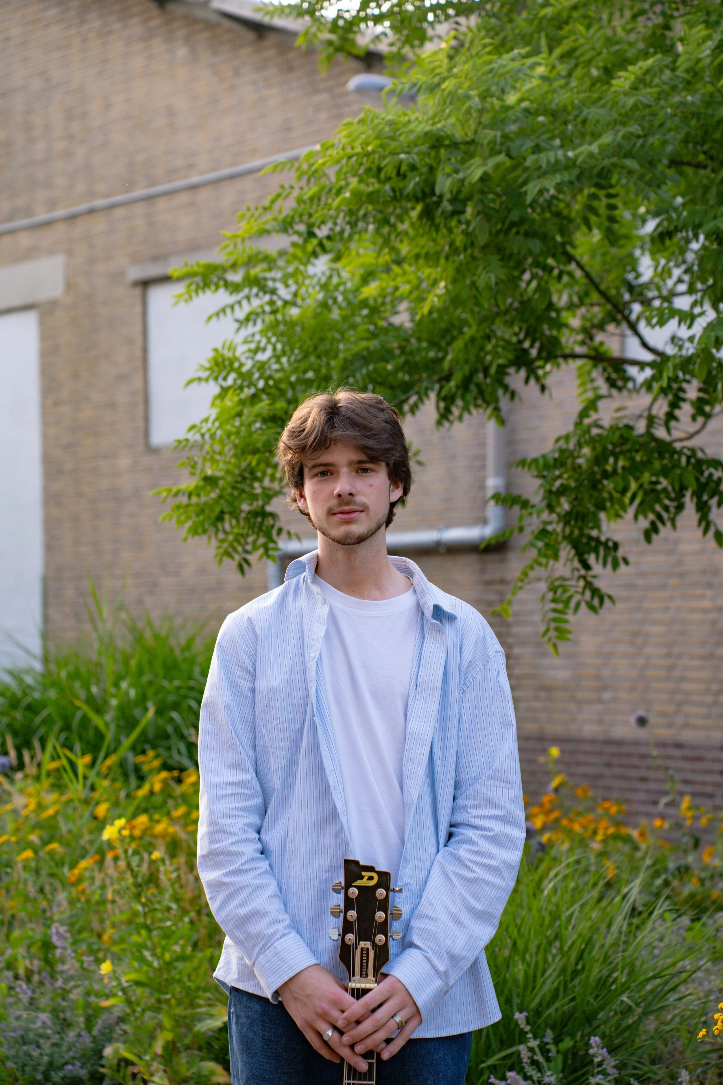 Young man with brown hair and beard standing outdoors in a garden, holding a guitar, wearing a blue and white striped shirt over a white T-shirt, with greenery and a brick building in the background.