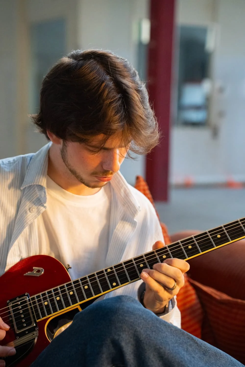 A young man with brown hair and a beard playing an electric guitar, sitting indoors on a red couch, with a background of a blurred room.