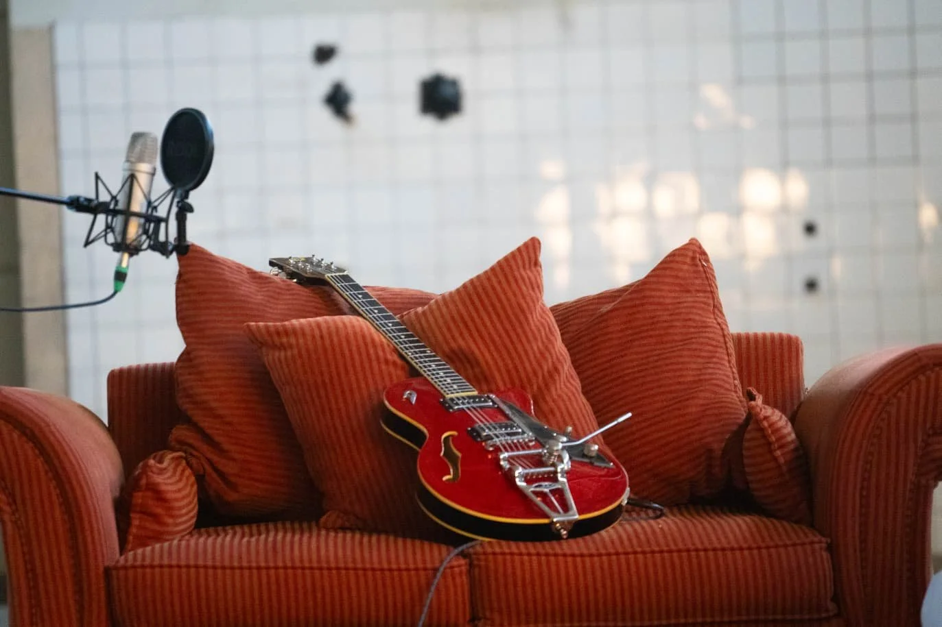 A red electric guitar resting on an orange striped couch with four cushions, positioned near a microphone in a recording or music studio.