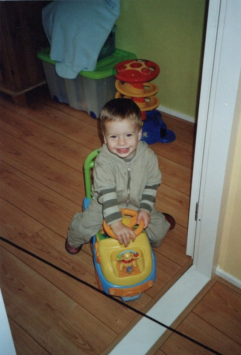 A young boy smiling and sitting on a toy car indoors, with wooden flooring and colorful toys in the background.