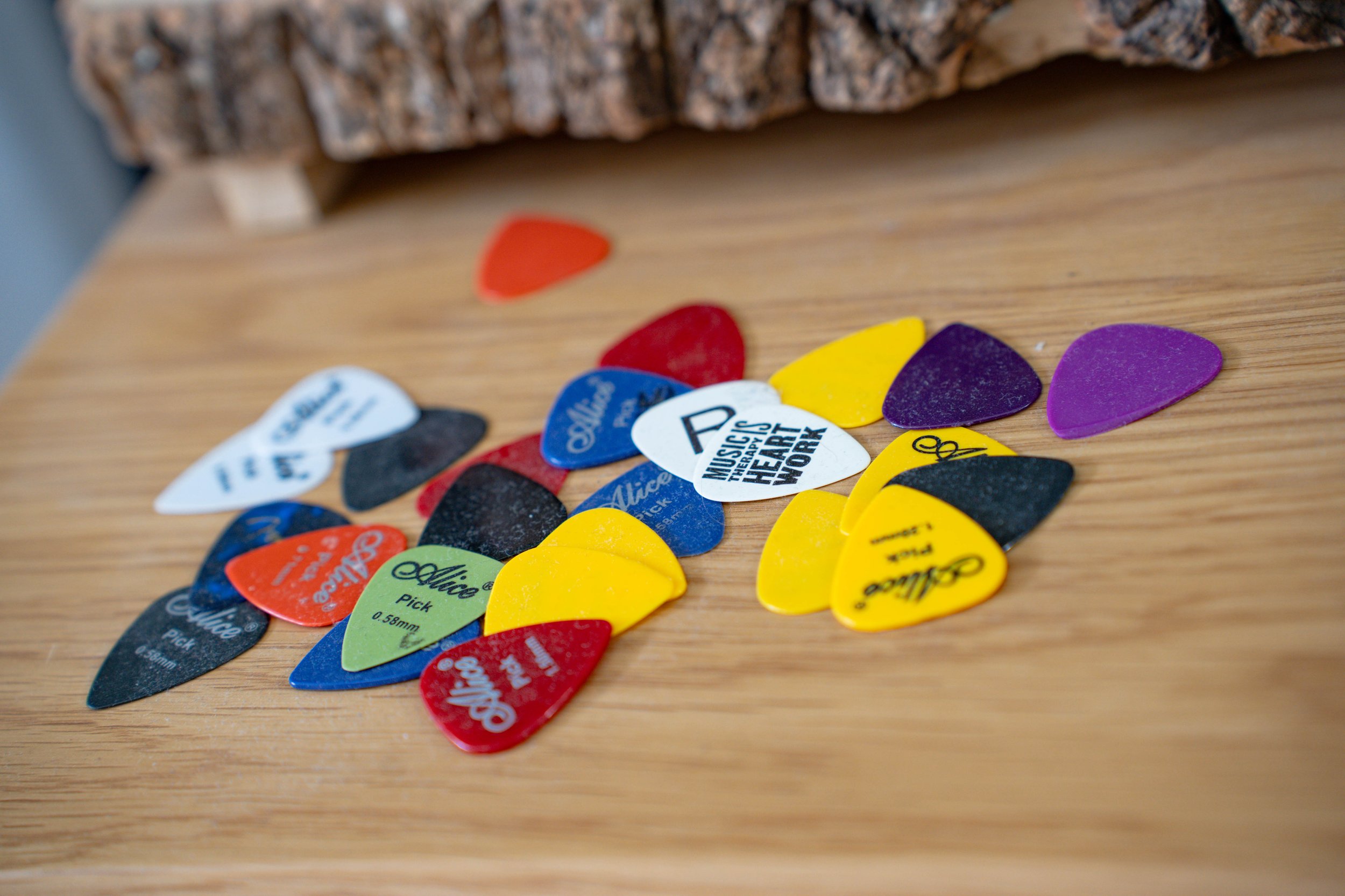 A variety of colorful guitar picks scattered on a wooden surface, with a rough-textured branch or piece of wood in the background.