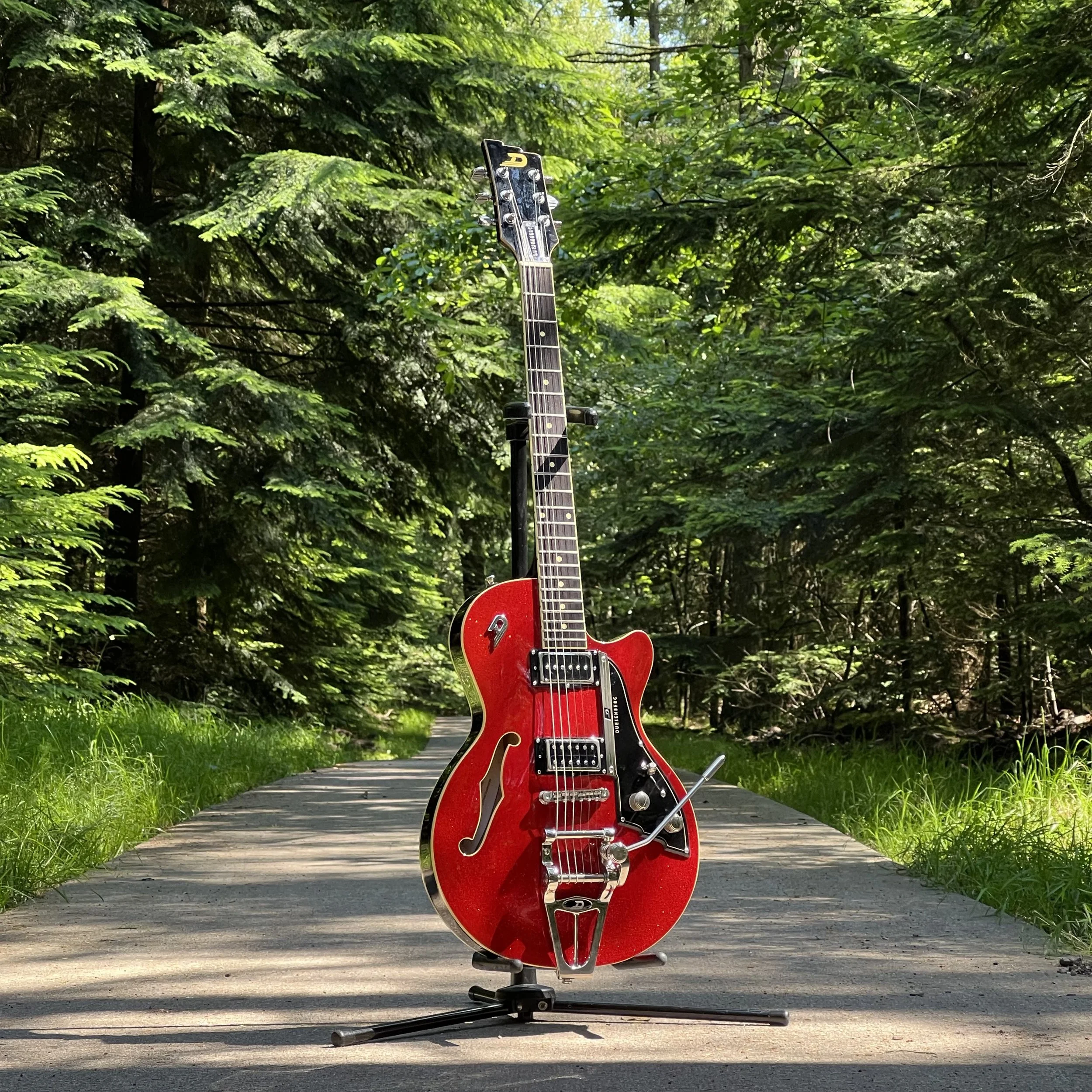 Red electric guitar standing on a guitar stand on a paved path surrounded by green trees and foliage.