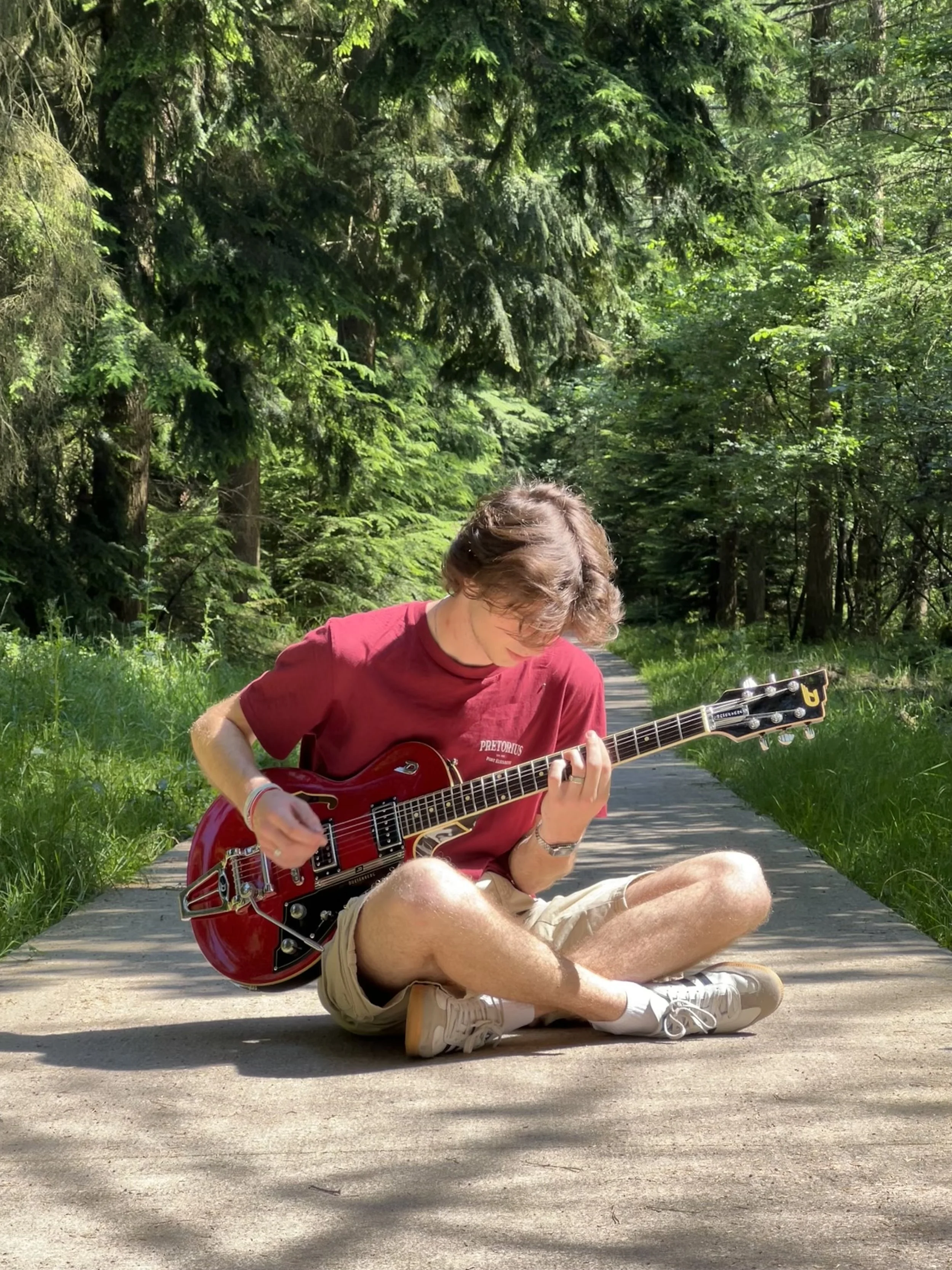 A person sitting on a sidewalk in a forested park, playing an electric guitar. The person is wearing a red shirt and beige shorts, with greenery and trees in the background.