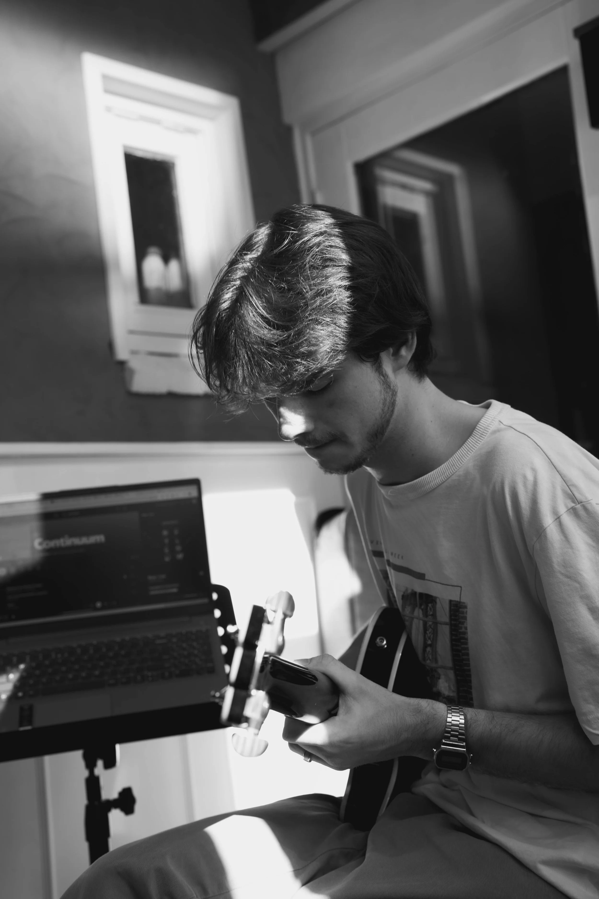 A young man with dark hair and a beard sitting in a room, looking at his smartphone, with a computer monitor in front of him, and a window with blinds in the background. The image is in black and white.