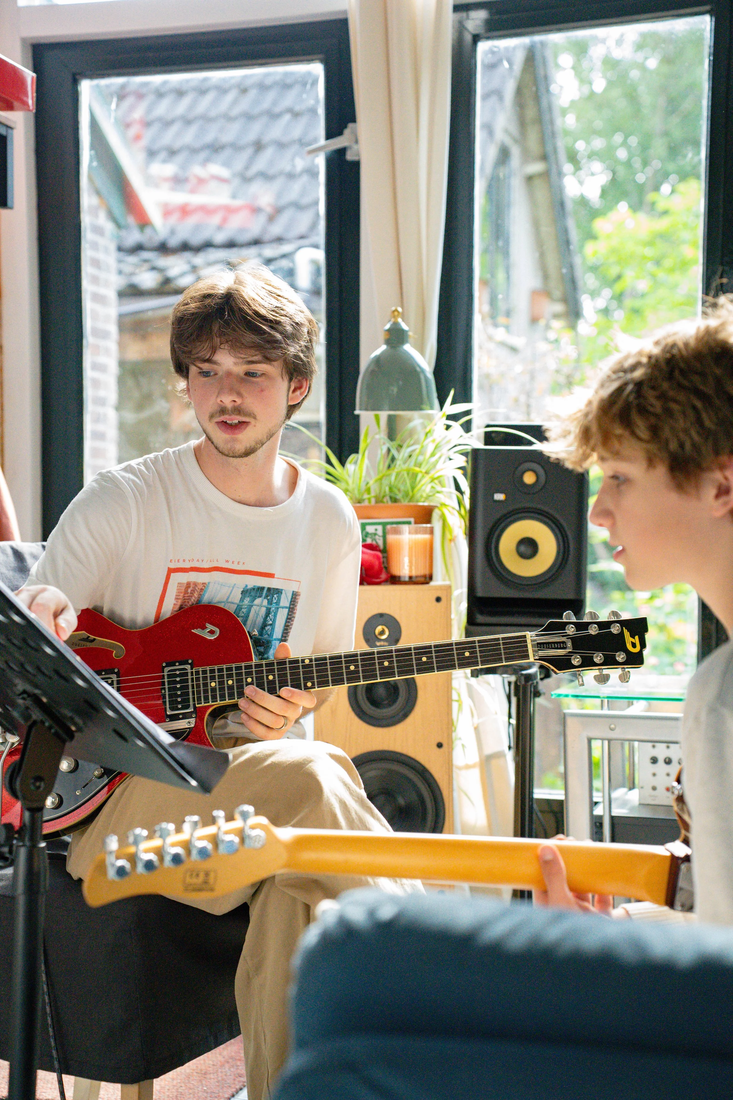 Two young men are playing guitars in a bright room with large windows, a plant, and audio equipment visible in the background.