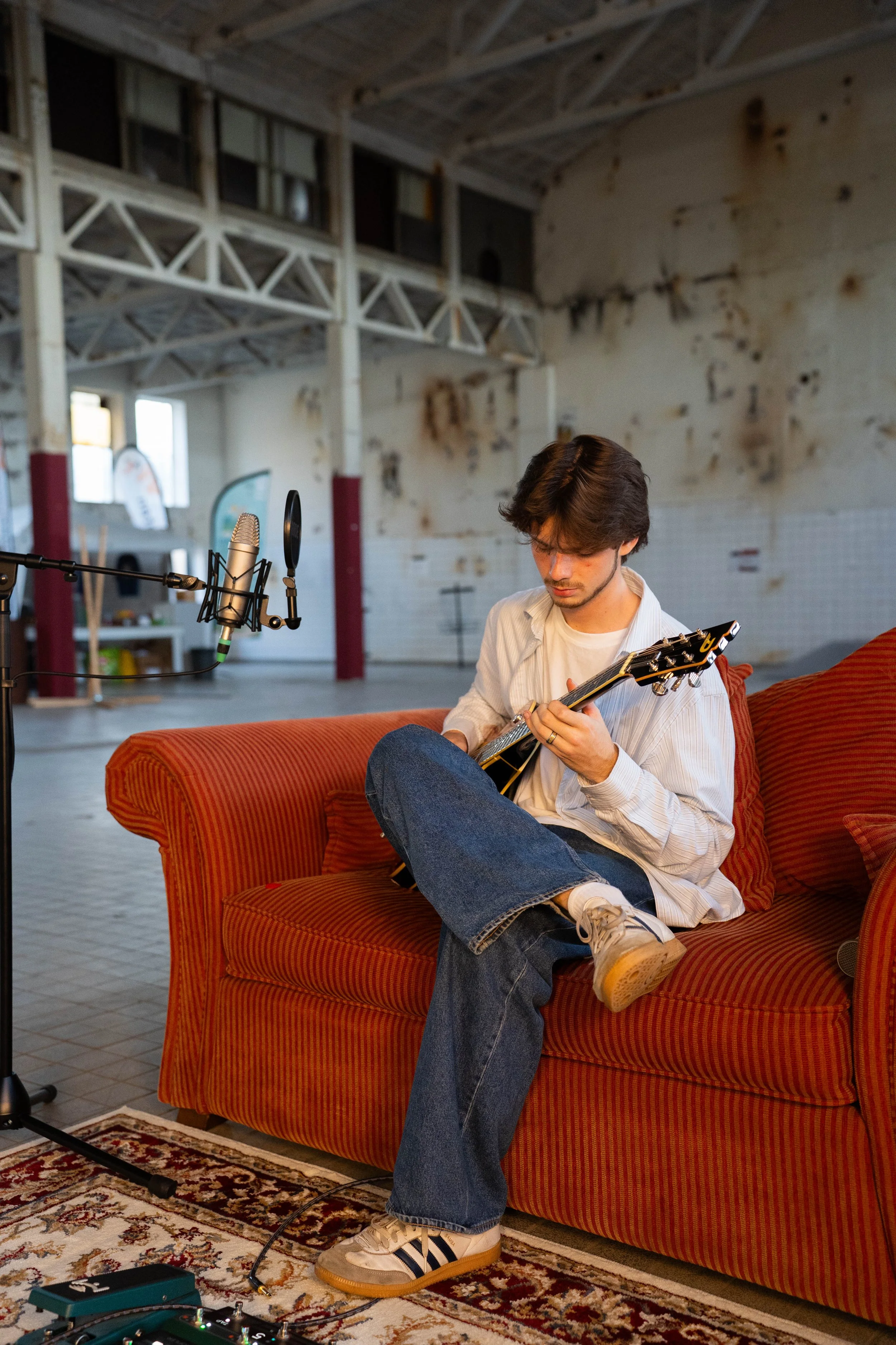 A young man with dark hair is sitting on an orange striped sofa in an industrial-style space, playing an electric guitar with a capo on the neck. A microphone on a stand is positioned in front of him. In the foreground, there is audio equipment on a patterned rug. The background features a high ceiling with exposed beams, a wall with peeling paint, and some objects on shelves.