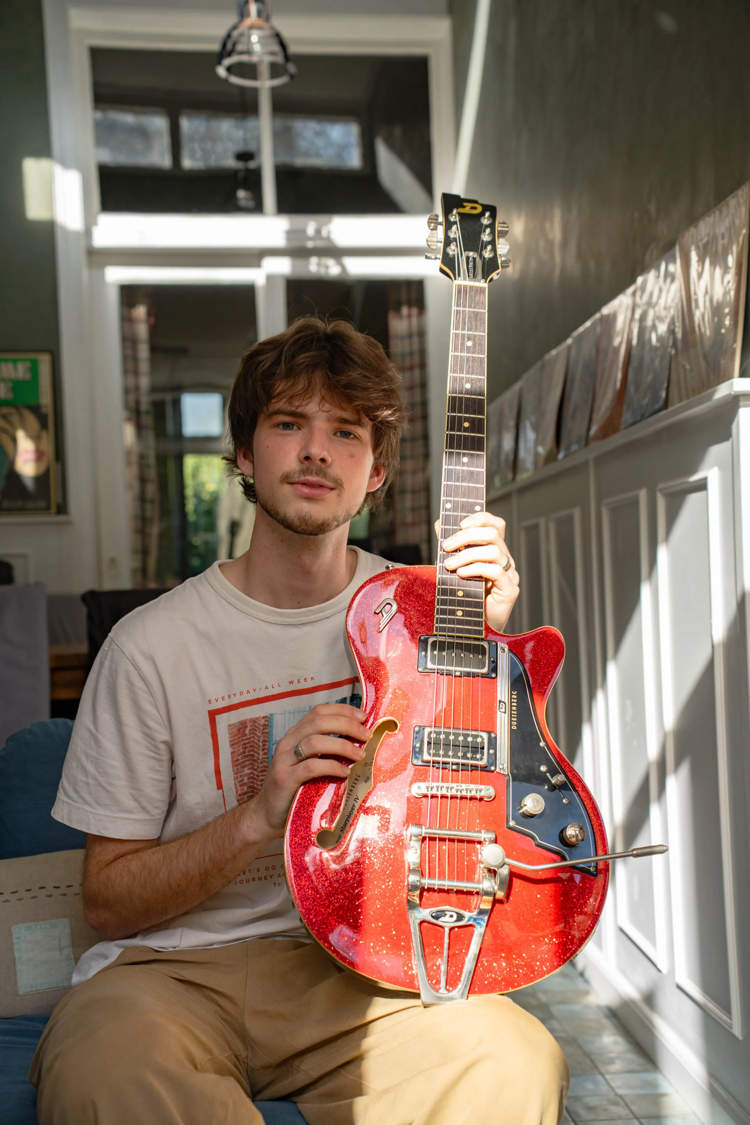 A young man with brown hair and a light beard sitting indoors, holding a red electric guitar with a distressed finish, in front of a window with curtains and a bookshelf.