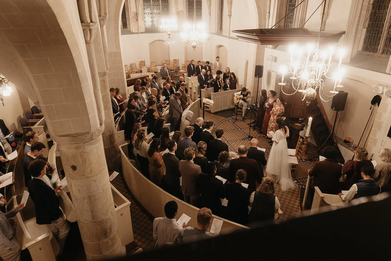 A congregation of people gathered inside a church for a ceremony, with some standing and others sitting, with a choir and musicians at the front near a piano and a large chandelier hanging from the ceiling.