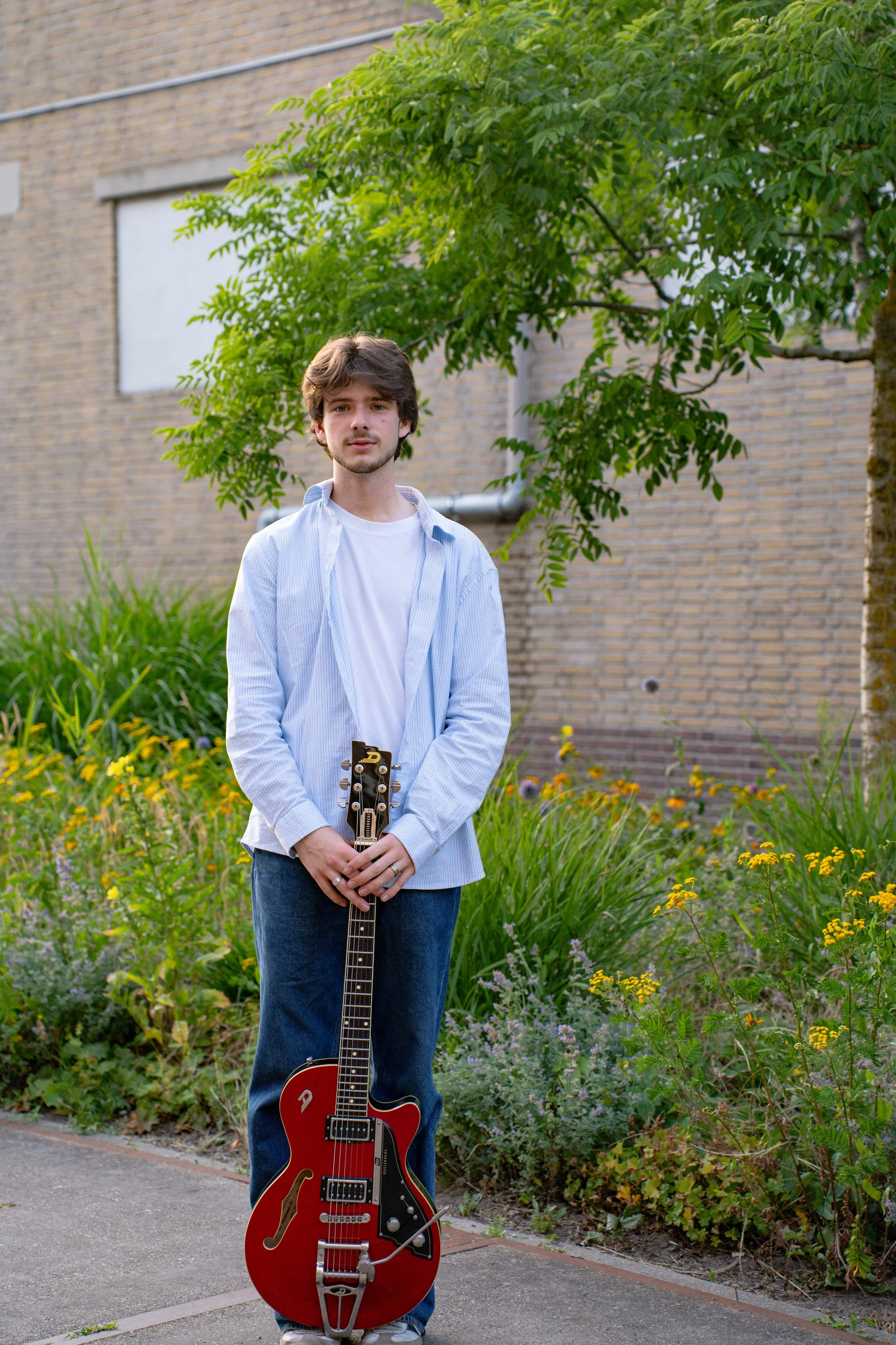 A young man with brown hair and light skin standing outdoors on a sidewalk, holding a red and black electric guitar. He is wearing a white t-shirt, a light blue button-up shirt, and jeans. Behind him is a lush green garden with various plants and flo
