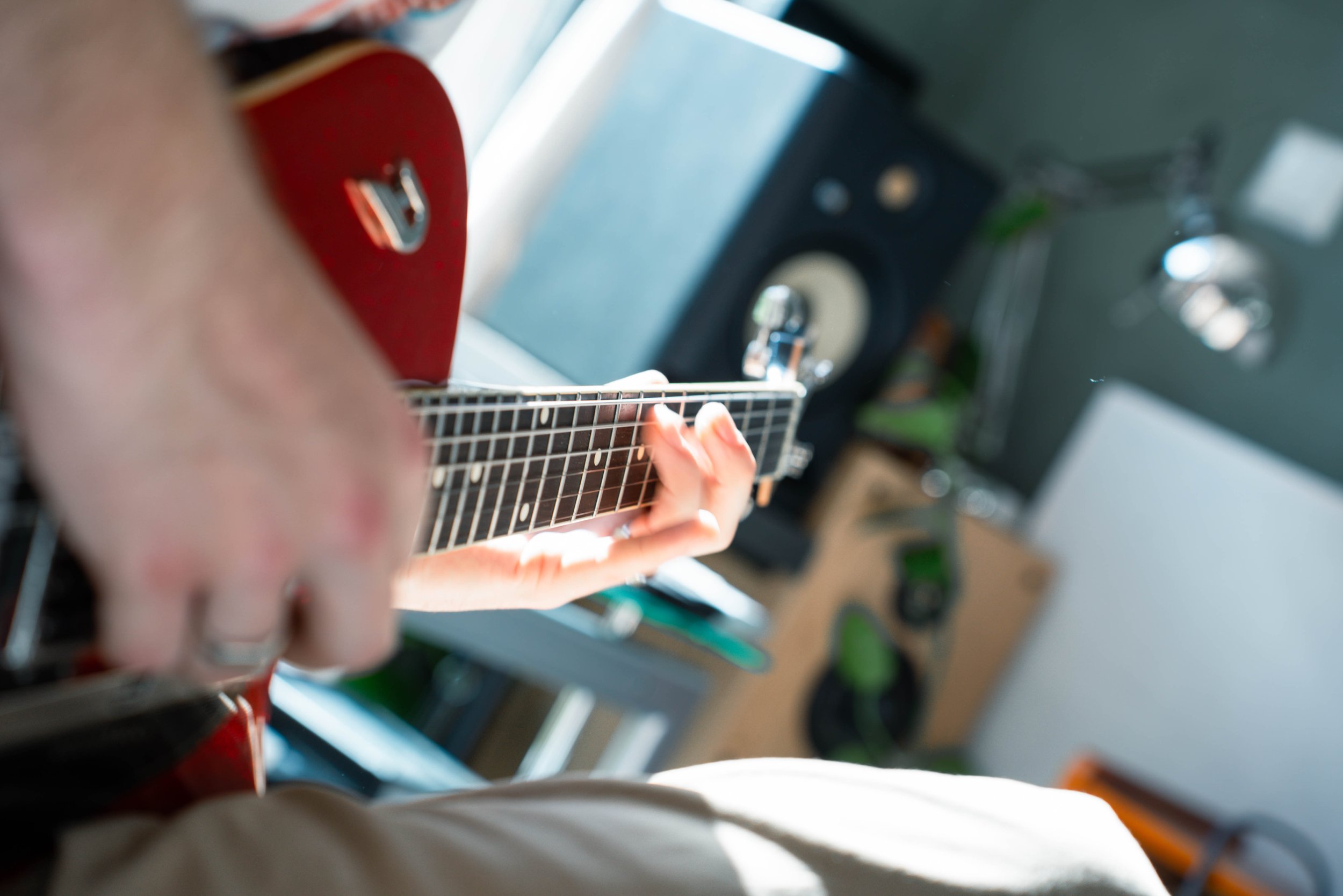 Close-up of a person's hand playing an acoustic guitar, with a speaker and other objects in the background.