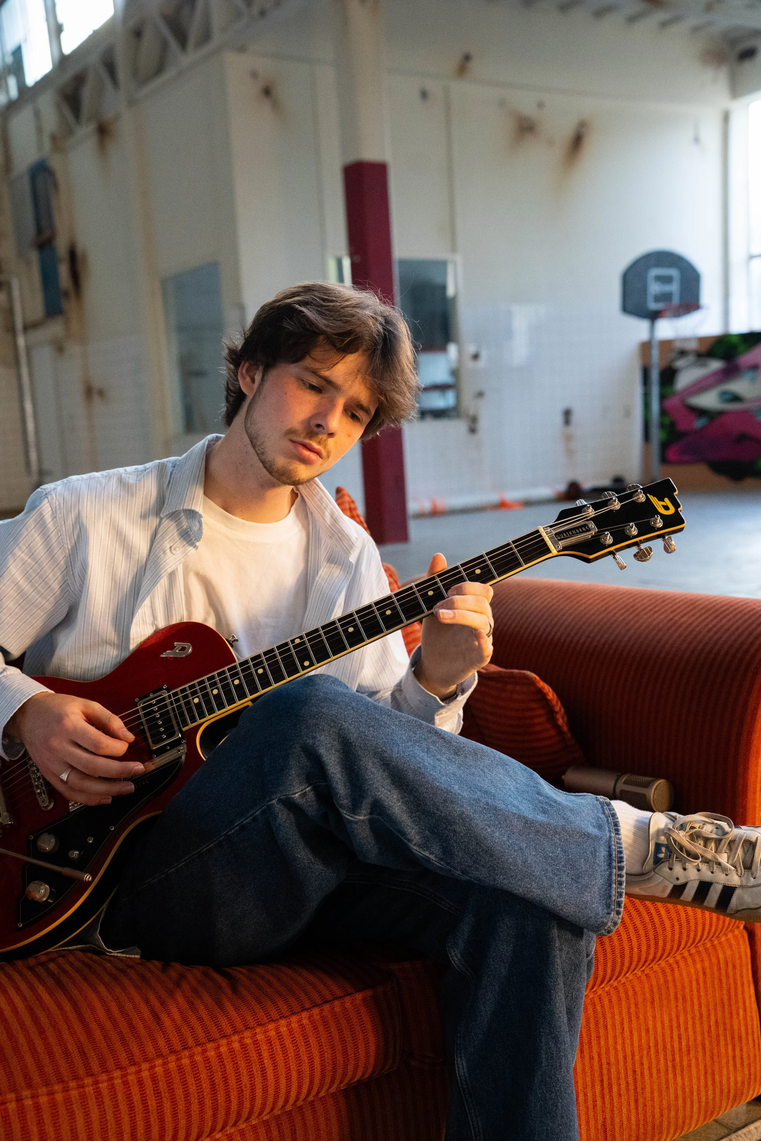 A young man with shoulder-length brown hair and a light beard, sitting on a red-orange striped couch in an indoor gymnasium or recreational area, playing a red electric guitar, wearing a white t-shirt, a light striped button-up shirt, and blue jeans.