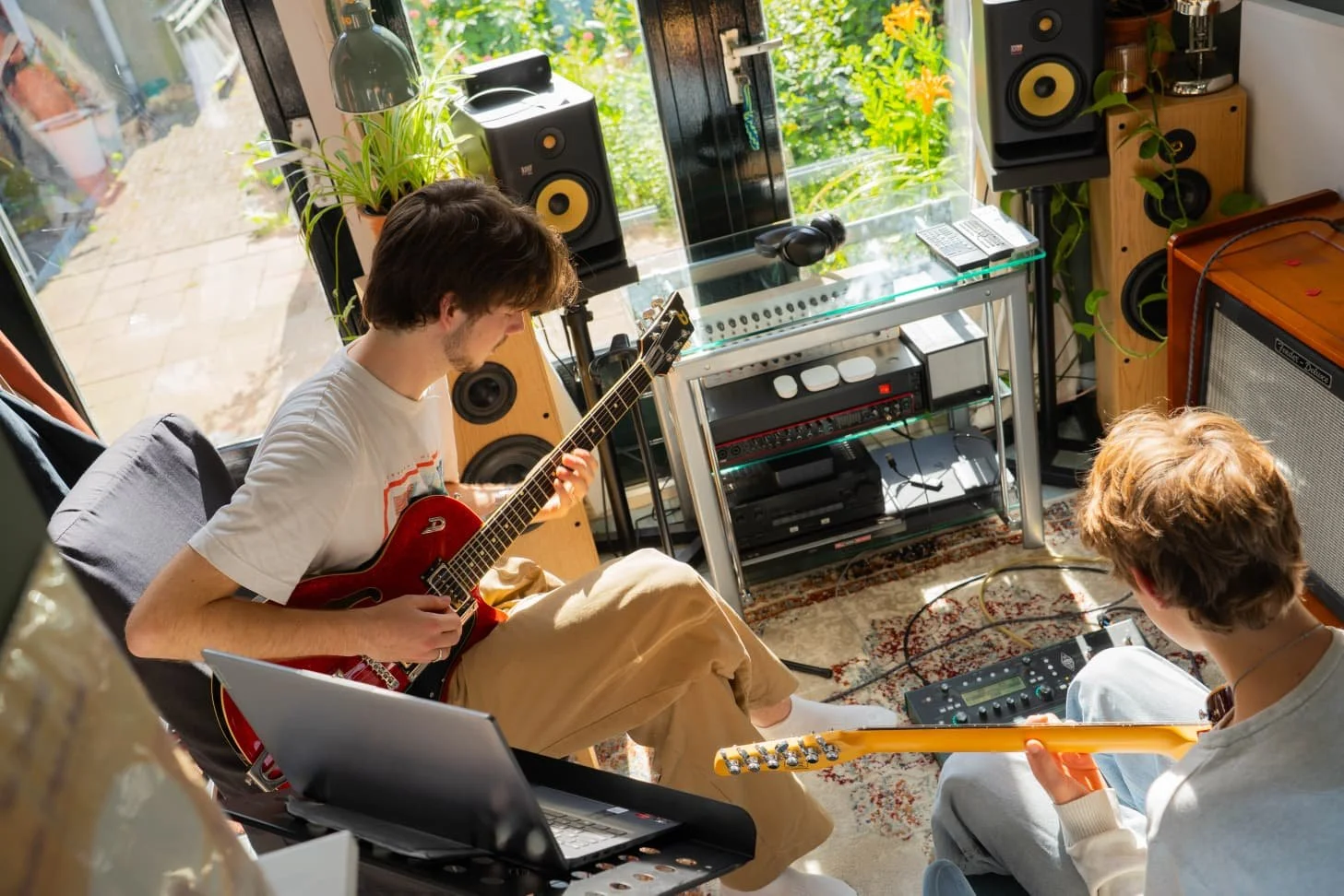 Two young men in a music studio practicing guitar, with one playing an electric guitar and the other an acoustic guitar, surrounded by audio equipment and speakers, in a well-lit room with large windows and plants.