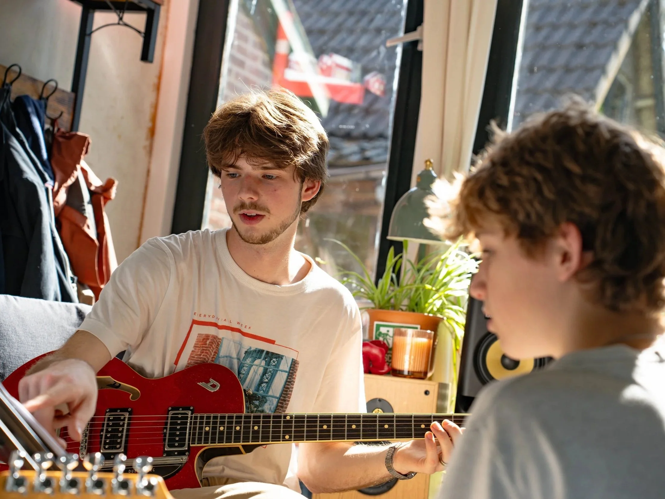 A young man with brown hair, wearing a white t-shirt, playing a red guitar. He is indoors near a large window with sunlight coming in, and there is a child with curly hair in the foreground. The background shows a coat rack with jackets and a view of rooftops outside.