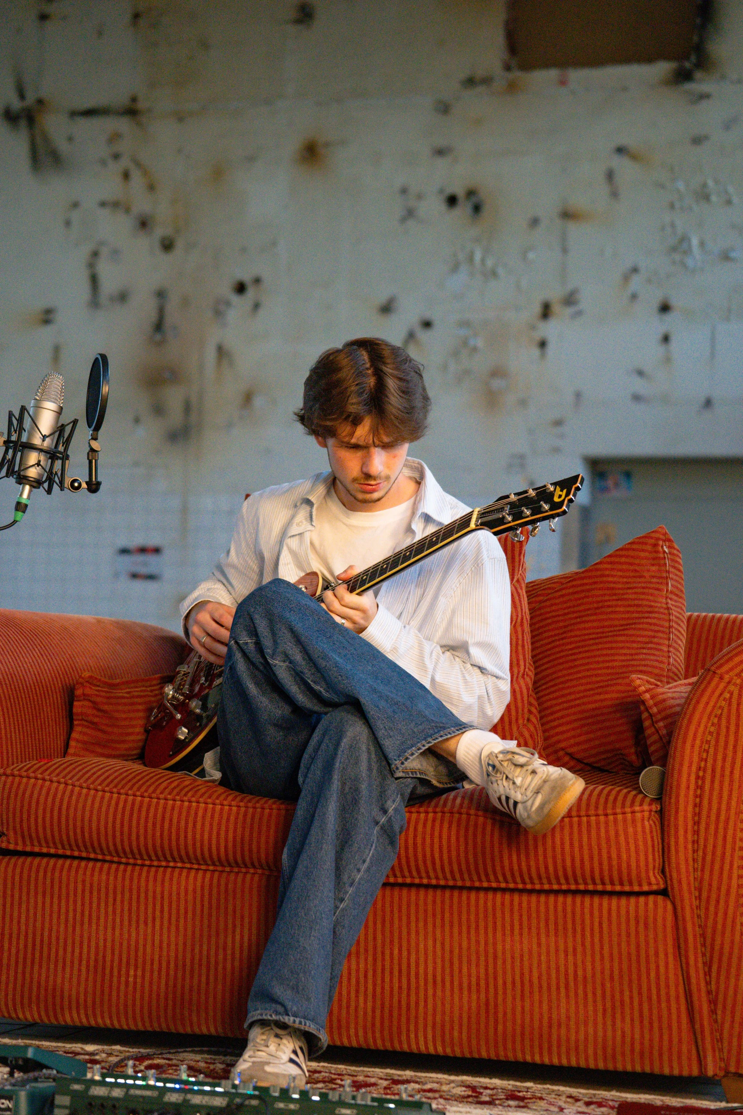 A young man with brown hair is sitting on an orange and red striped couch, playing a guitar. He is wearing a white shirt, blue jeans, and white sneakers. There is a microphone on a stand and audio equipment visible nearby. The background features a p