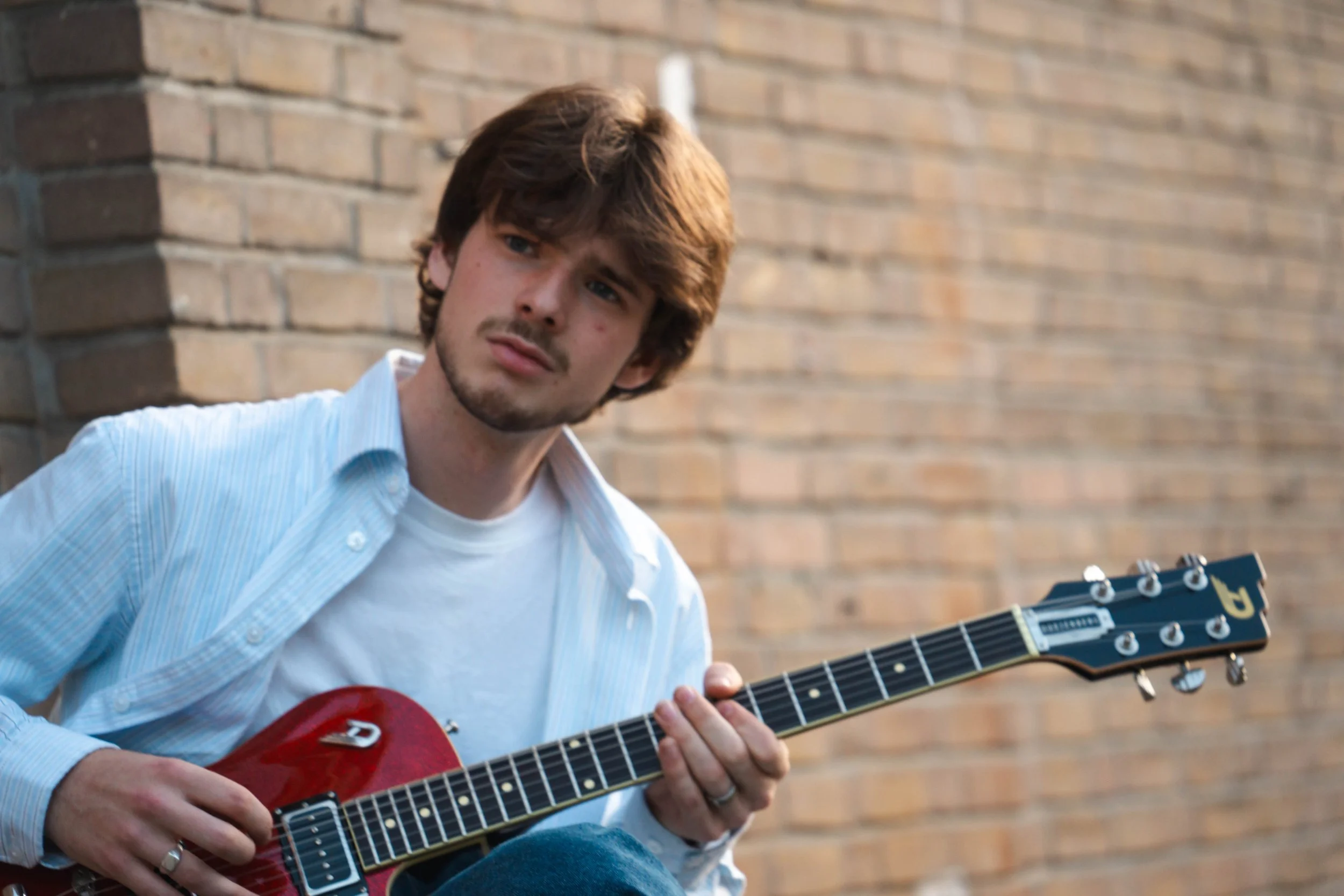 Young man with brown hair and a mustache playing a red electric guitar in front of a brick wall.