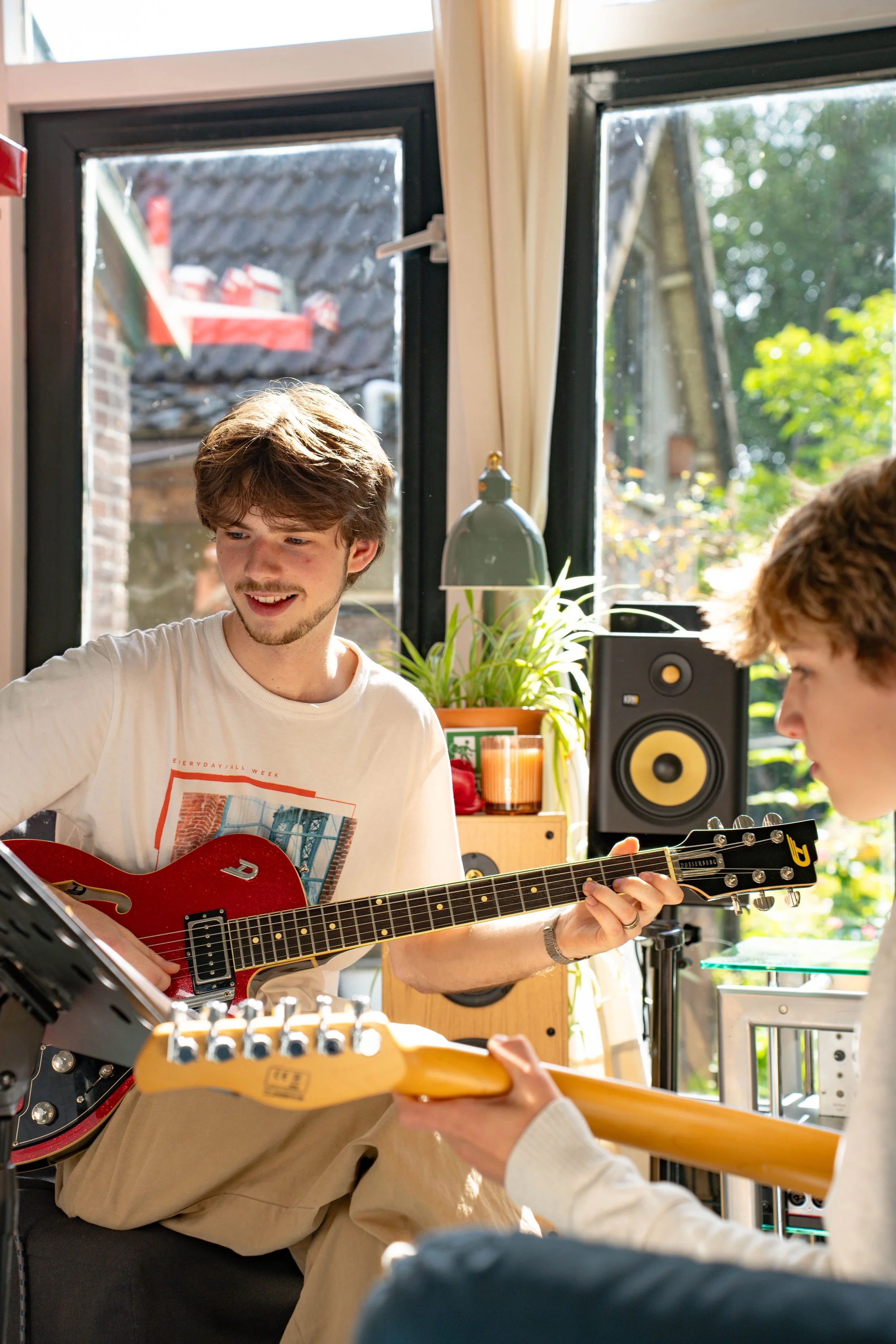 Two young men playing electric guitars and a third person holding a bass guitar in a sunlit room with large windows and a plant.