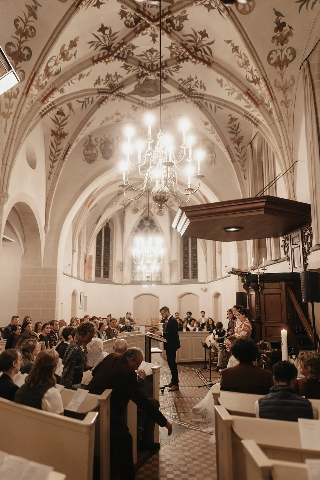 People attending a service or ceremony in a church with high vaulted, decorated ceilings and chandeliers, and a man speaking at a lectern.