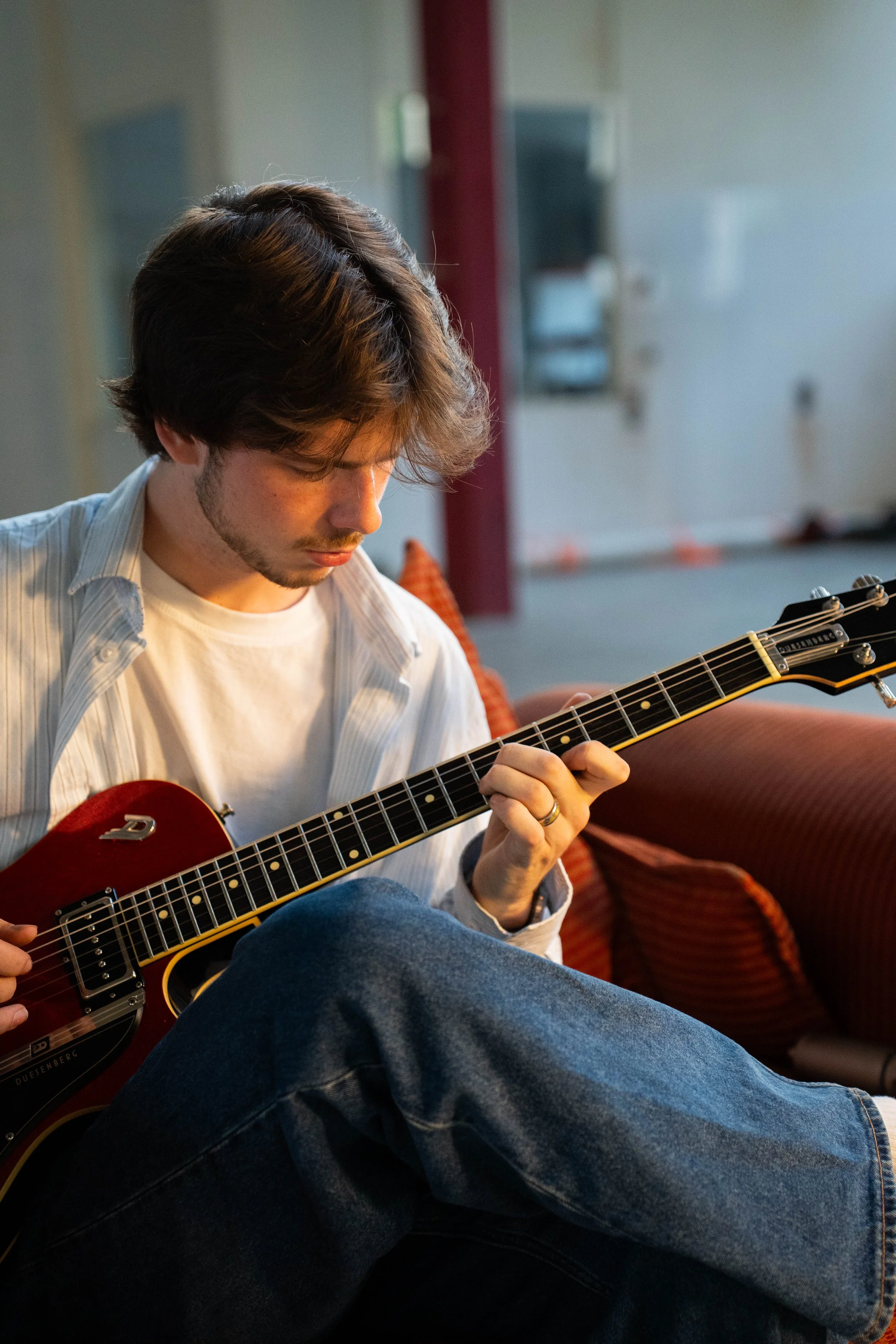 A young man with brown hair and a beard is sitting on a red couch, playing an electric guitar in a room with a blurred background.