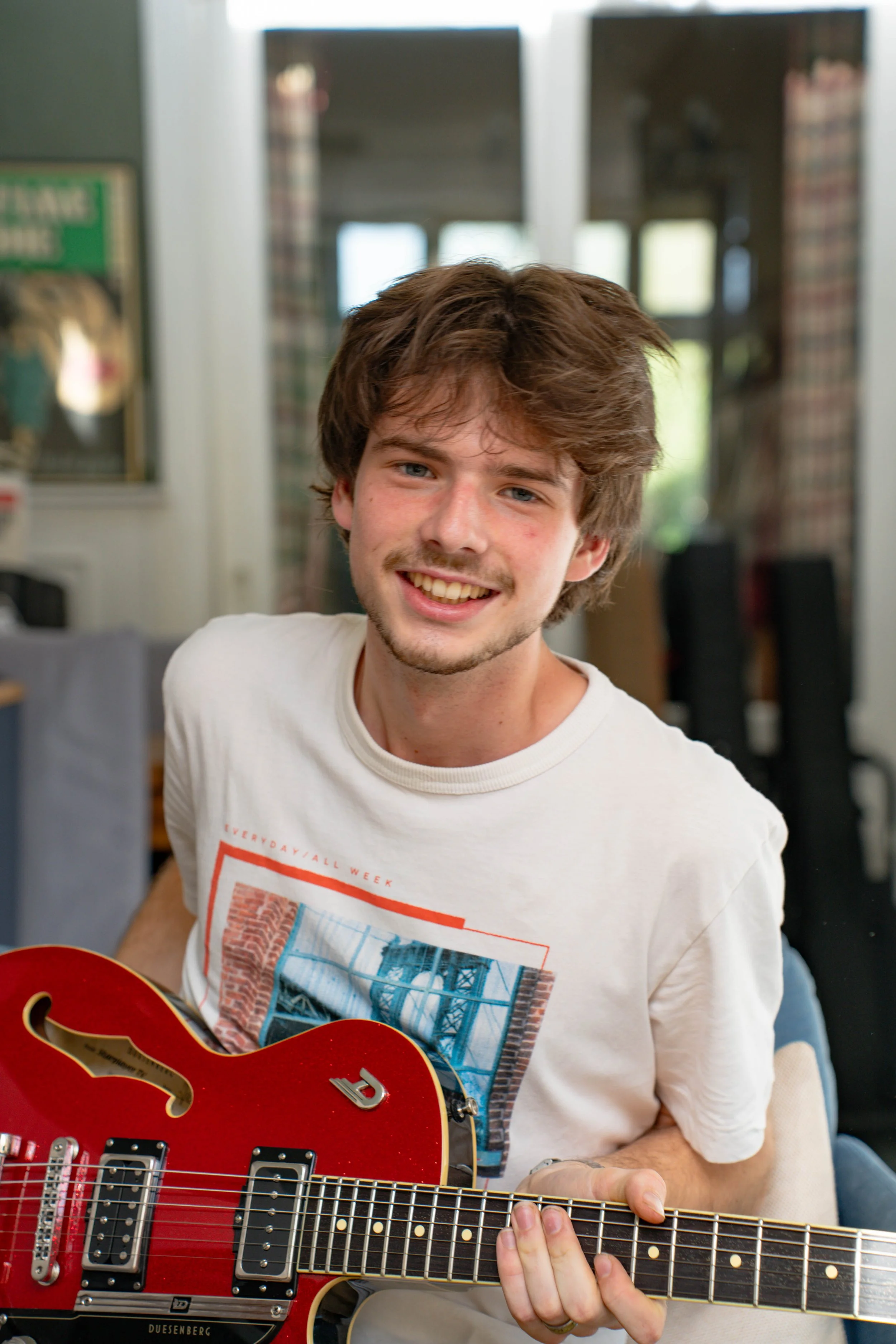 A young man with brown hair and a light beard playing a red electric guitar in a room with a blurred background of windows and plaid curtains.
