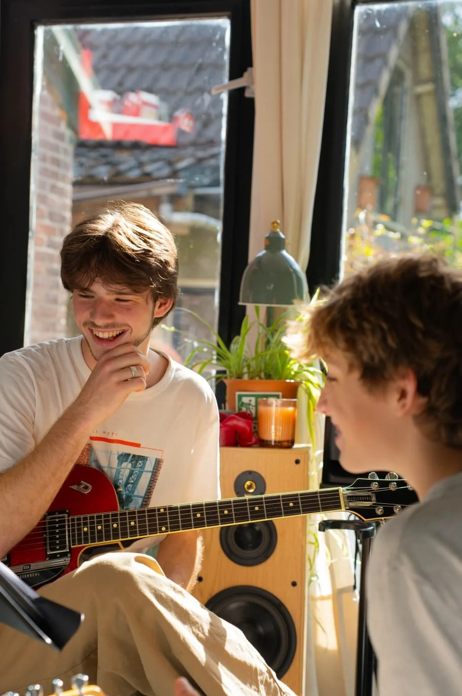 Two young men are enjoying music and each other’s company in a bright room. One is playing a red guitar while the other is smiling and leaning in, creating a joyful atmosphere.